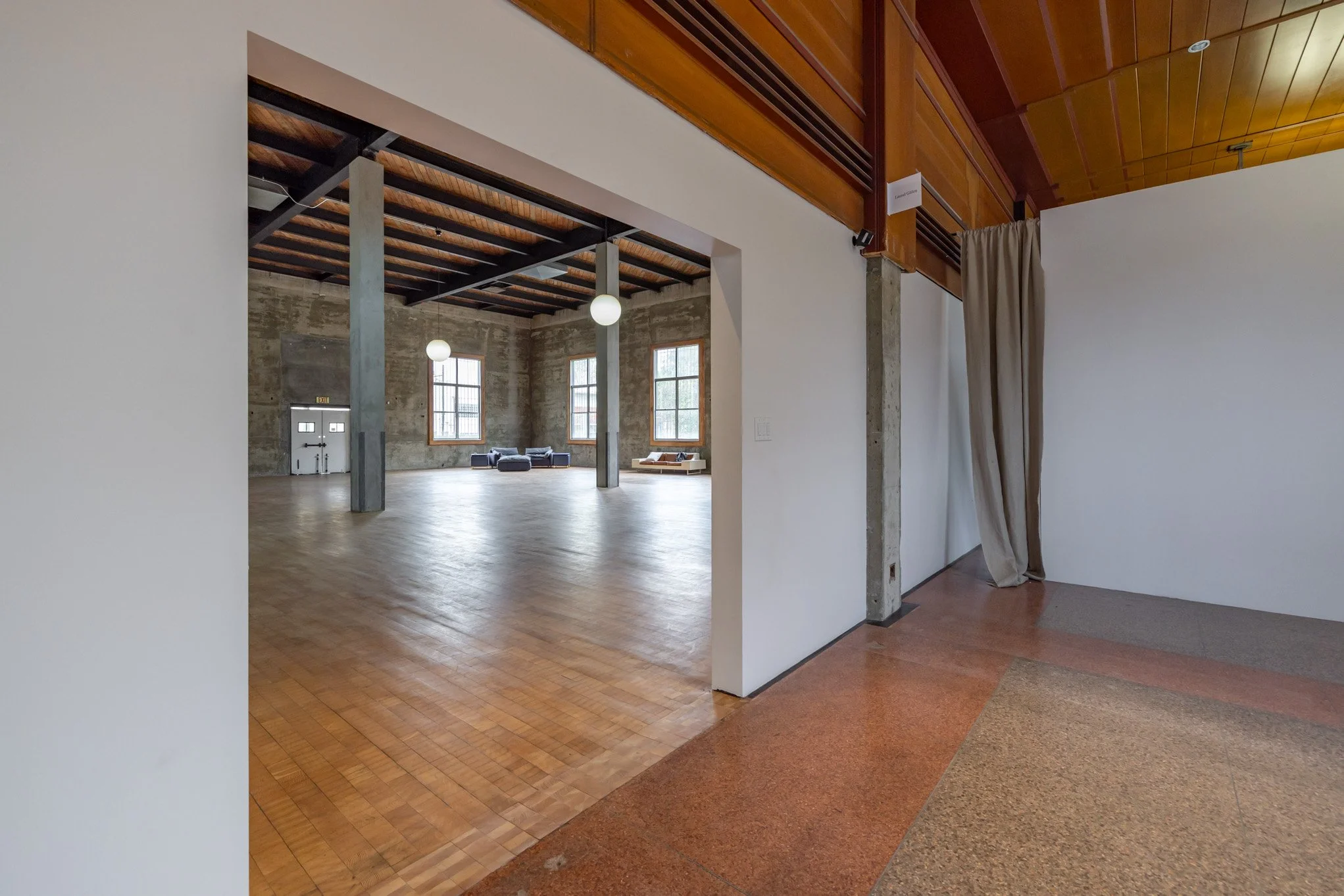 Interior of an industrial-style building with large windows, wooden floors and ceiling, and concrete support beams, seen from an adjacent white wall area with a curtain.