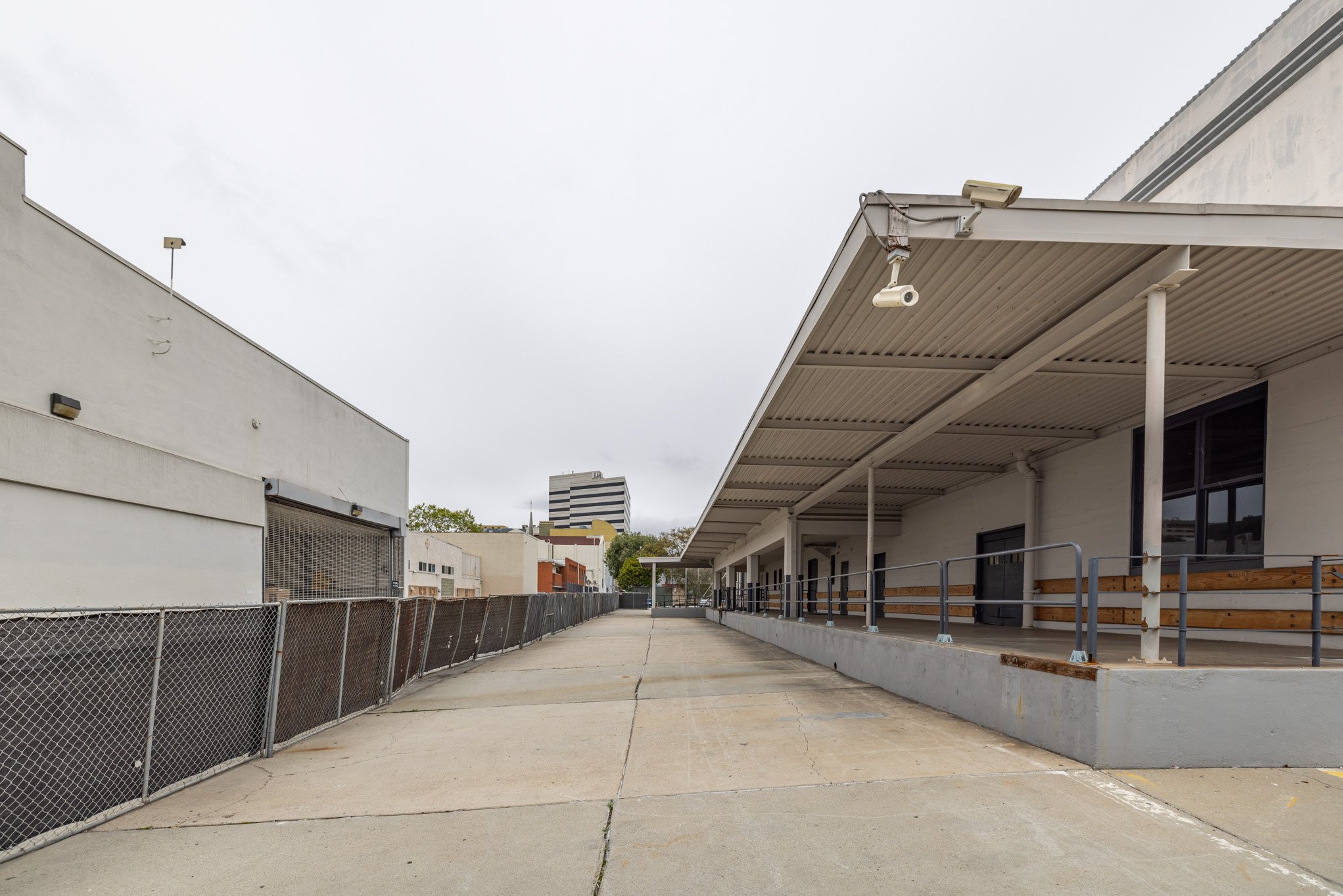 Empty outdoor alley with a covered sidewalk on the right side, white buildings, and a chain-link fence on the left, under an overcast sky.