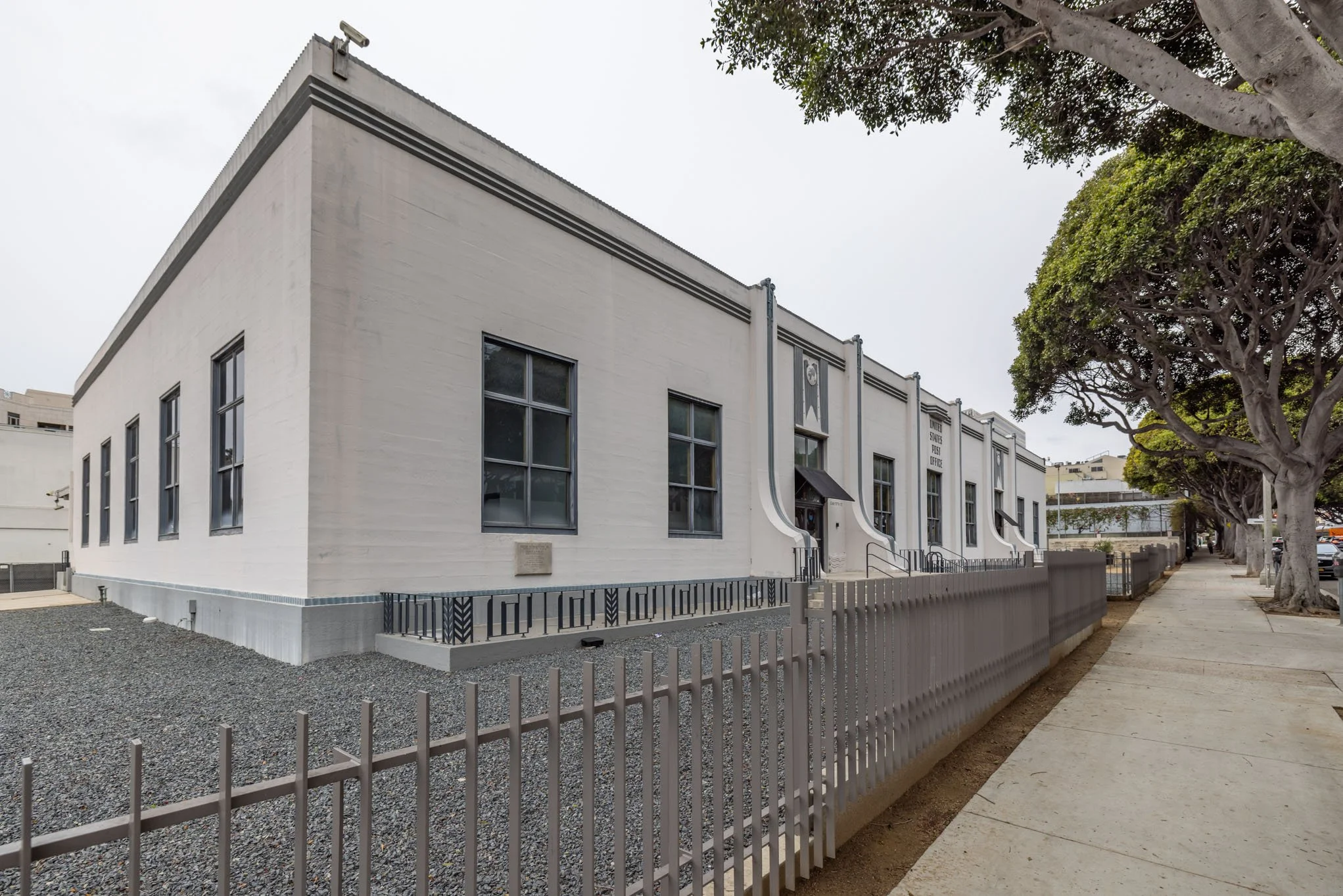 Front exterior of a white, two-story government building with tall windows, black railings, and a sidewalk with trees.