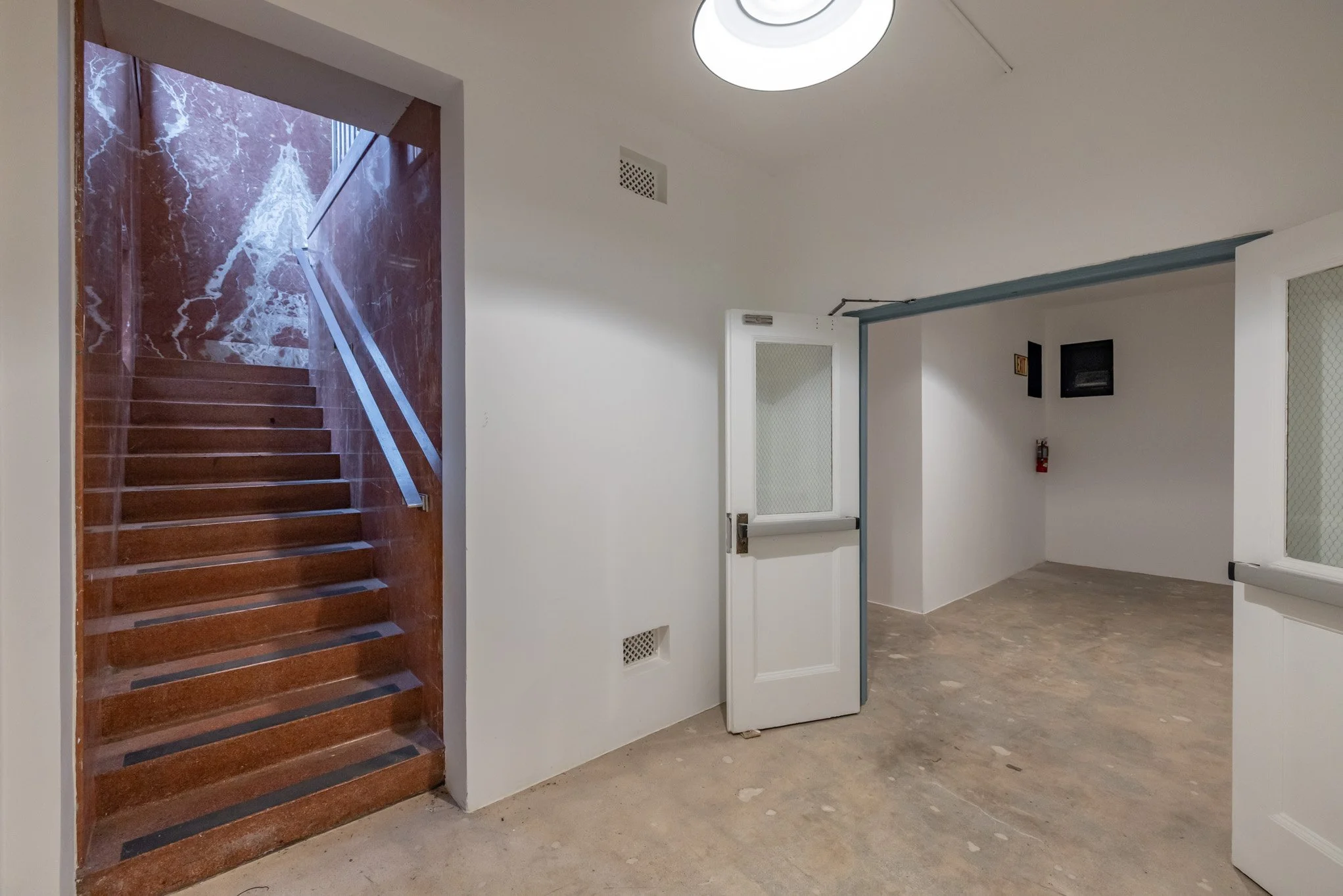 Empty indoor space with a staircase behind a glass door, featuring wooden steps and a metal handrail, and a white door with a glass window, in a room with white walls and ceiling.