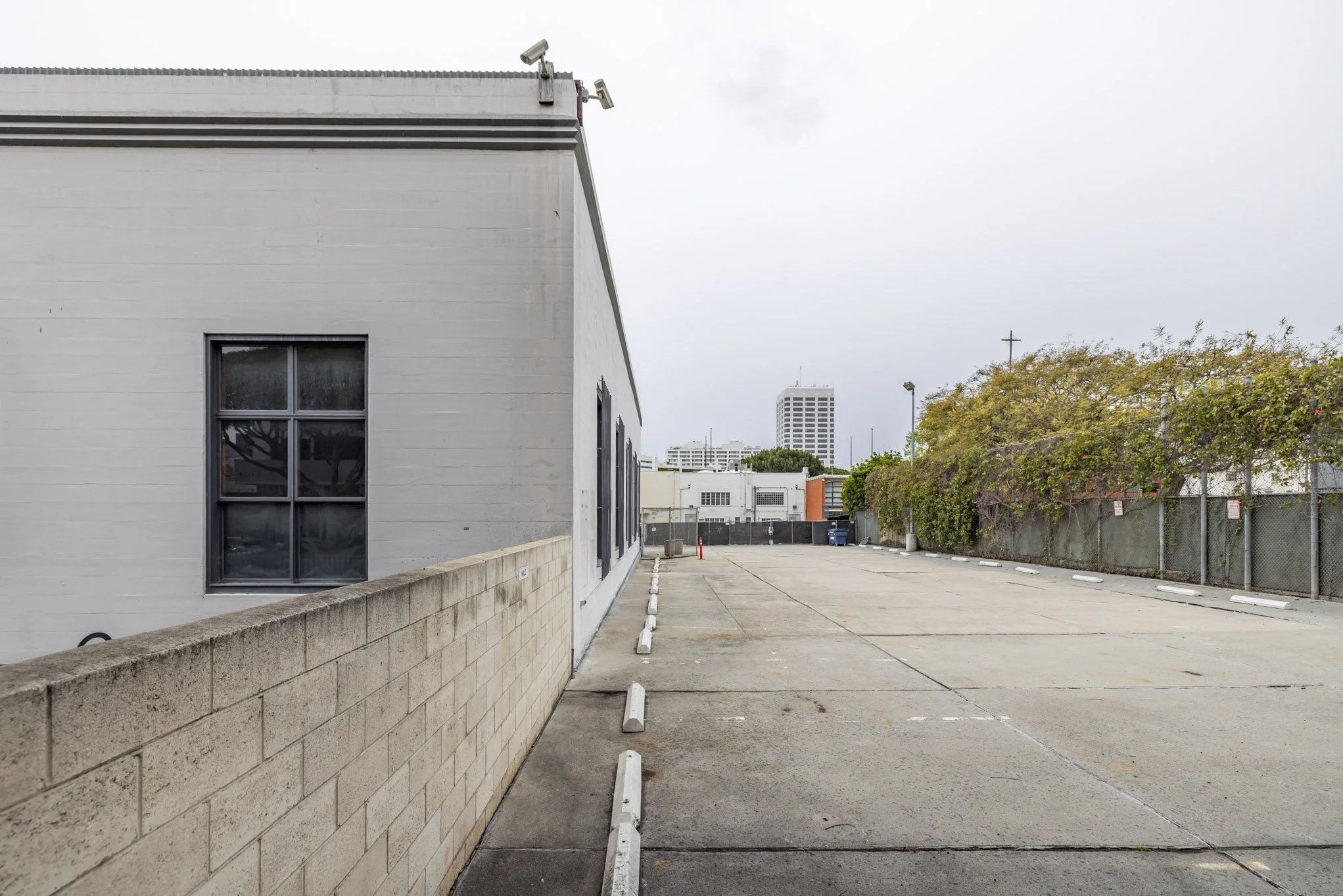 Empty parking lot next to a white building with windows, surrounded by a concrete wall and a chain-link fence covered in greenery, with city high-rise buildings in the background under an overcast sky.