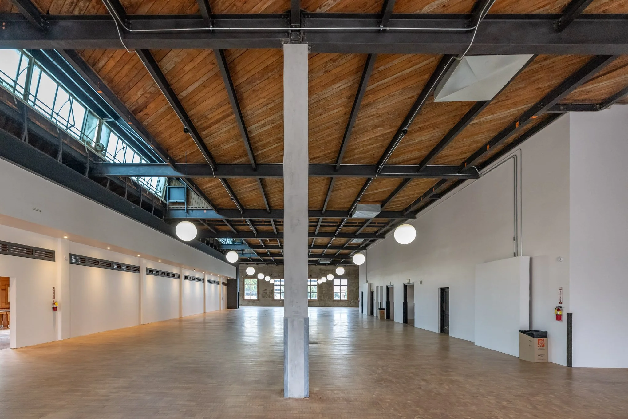 Empty open-concept room with hardwood floors, white walls, large windows, and a wooden ceiling with exposed beams. Decorated with round hanging lights and some doors along the right wall.