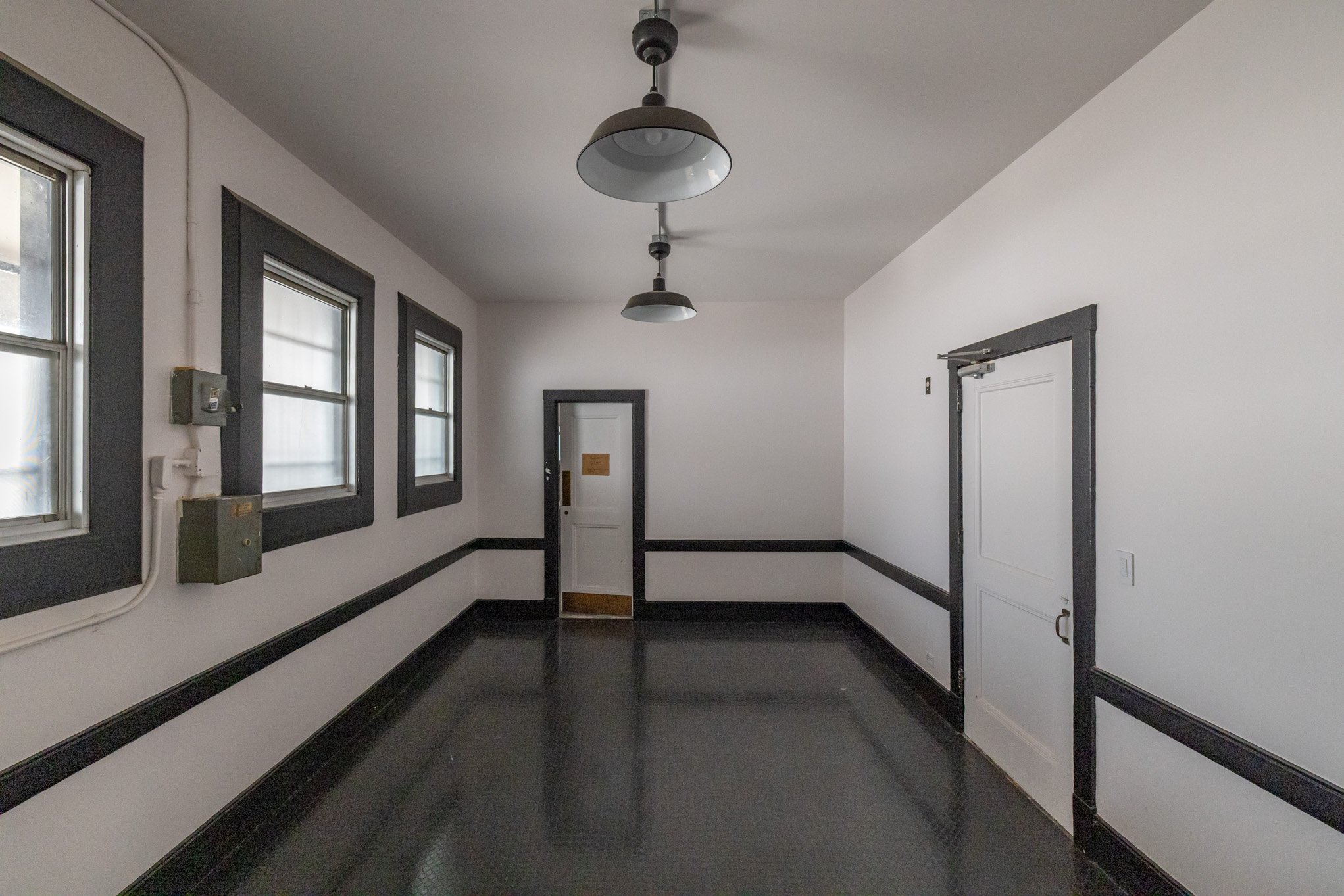 Empty room with white walls, three windows with black trim, black baseboards, and overhead lighting fixtures.