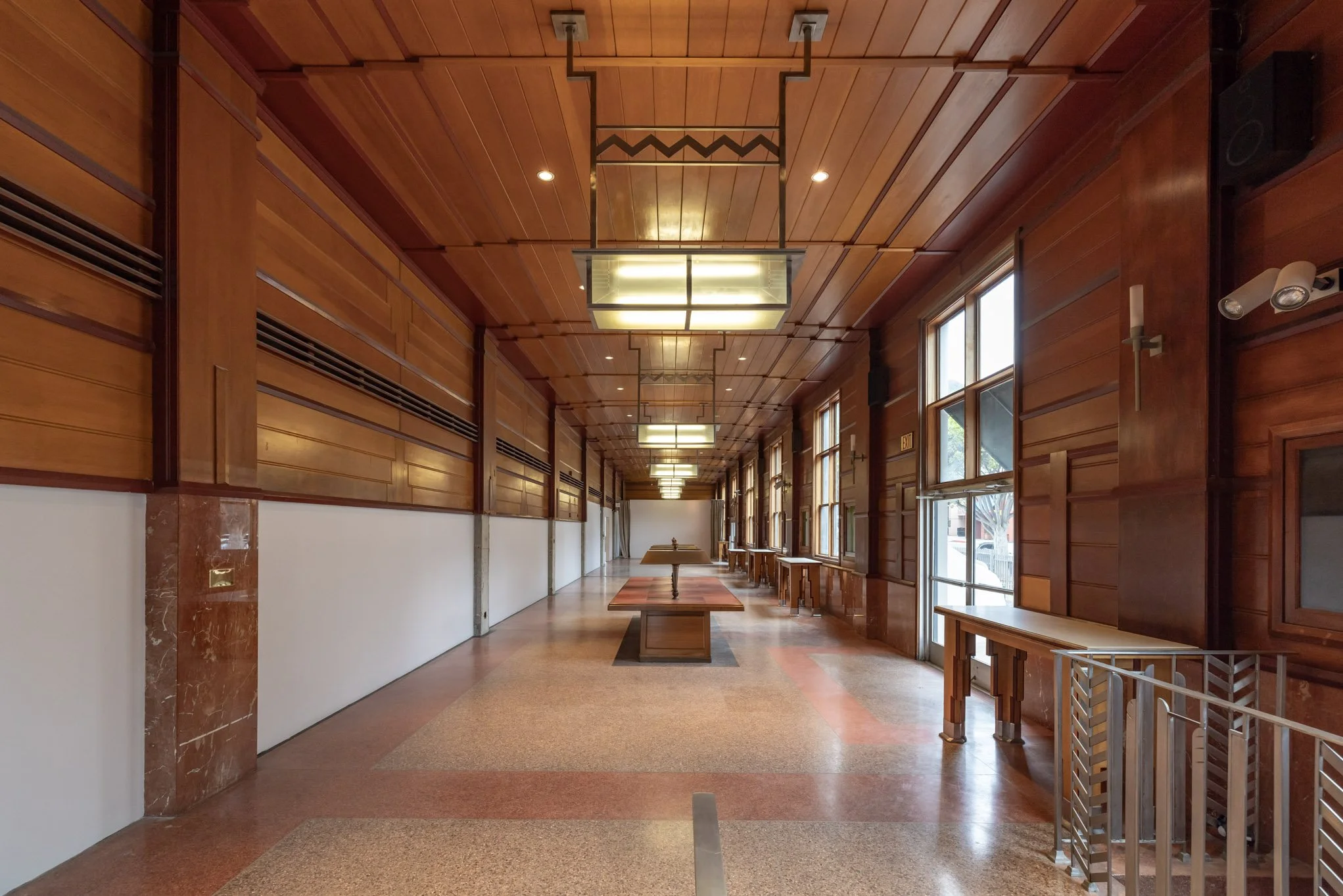 Long corridor with wooden paneling, large windows on the right, ceiling lights, tables along the right wall, and an empty wooden floor.