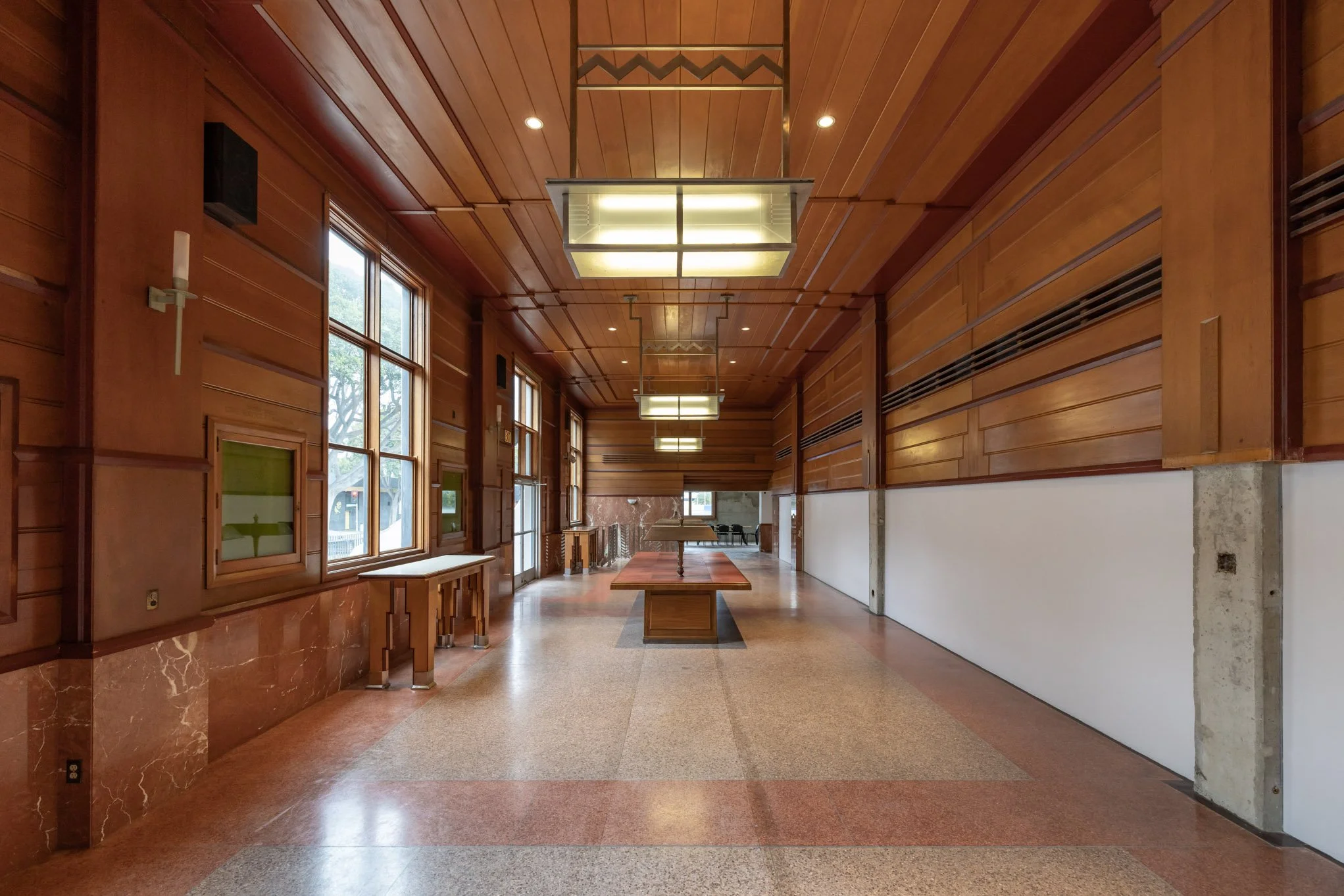 Long hall with wood-paneled walls, large windows, and rectangular light fixtures hanging from the ceiling. There are tables along the center and chairs at the far end.