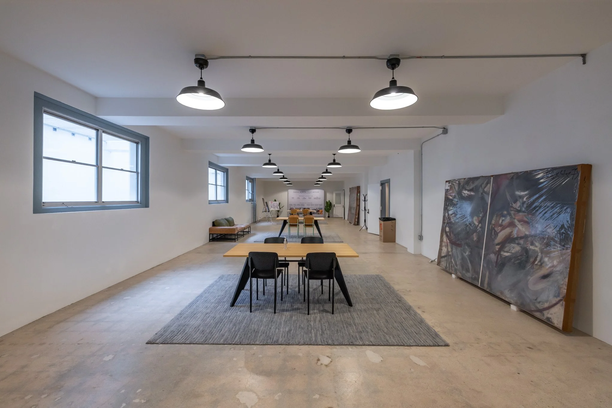 Spacious art gallery with white walls, large windows, modern black ceiling lights, minimalist furniture, and various paintings and artworks displayed