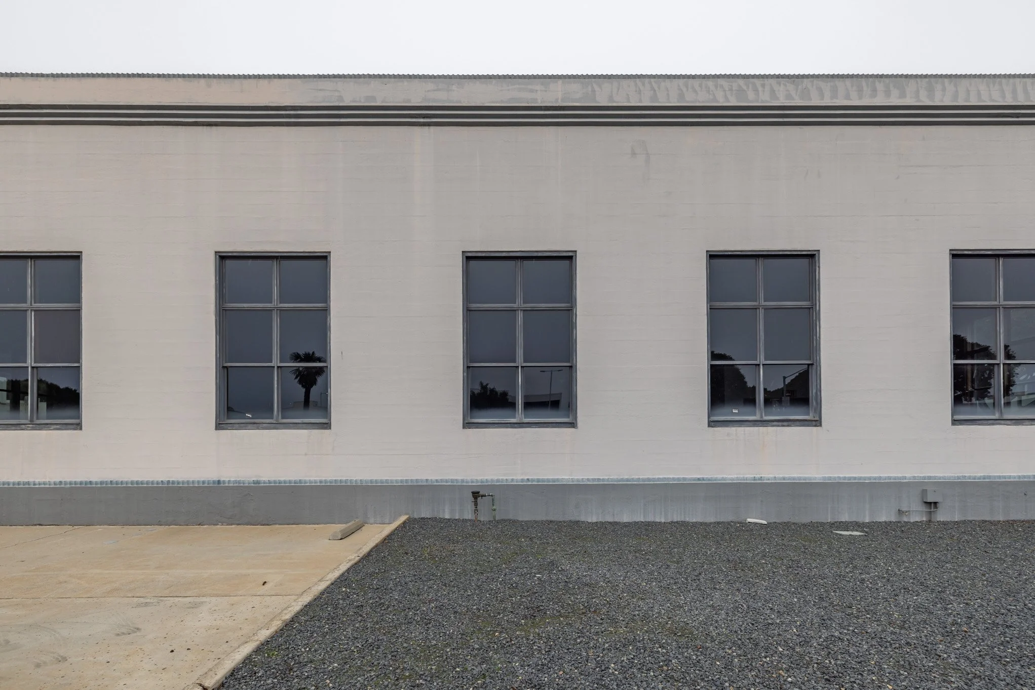 Exterior of a white brick building with four windows, two with reflections of palm trees, against a cloudy sky