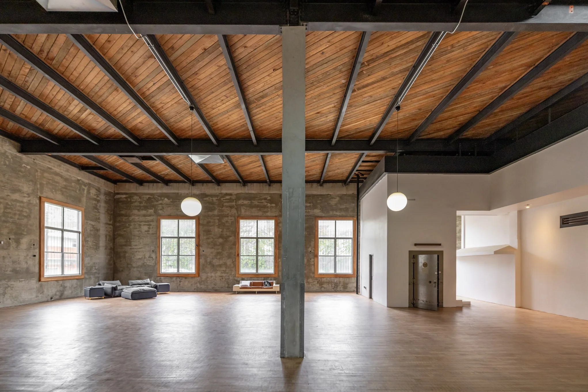 Spacious interior with exposed brick walls, wooden ceiling with black beams, large windows, and minimalist decor including a few sofas and side table.