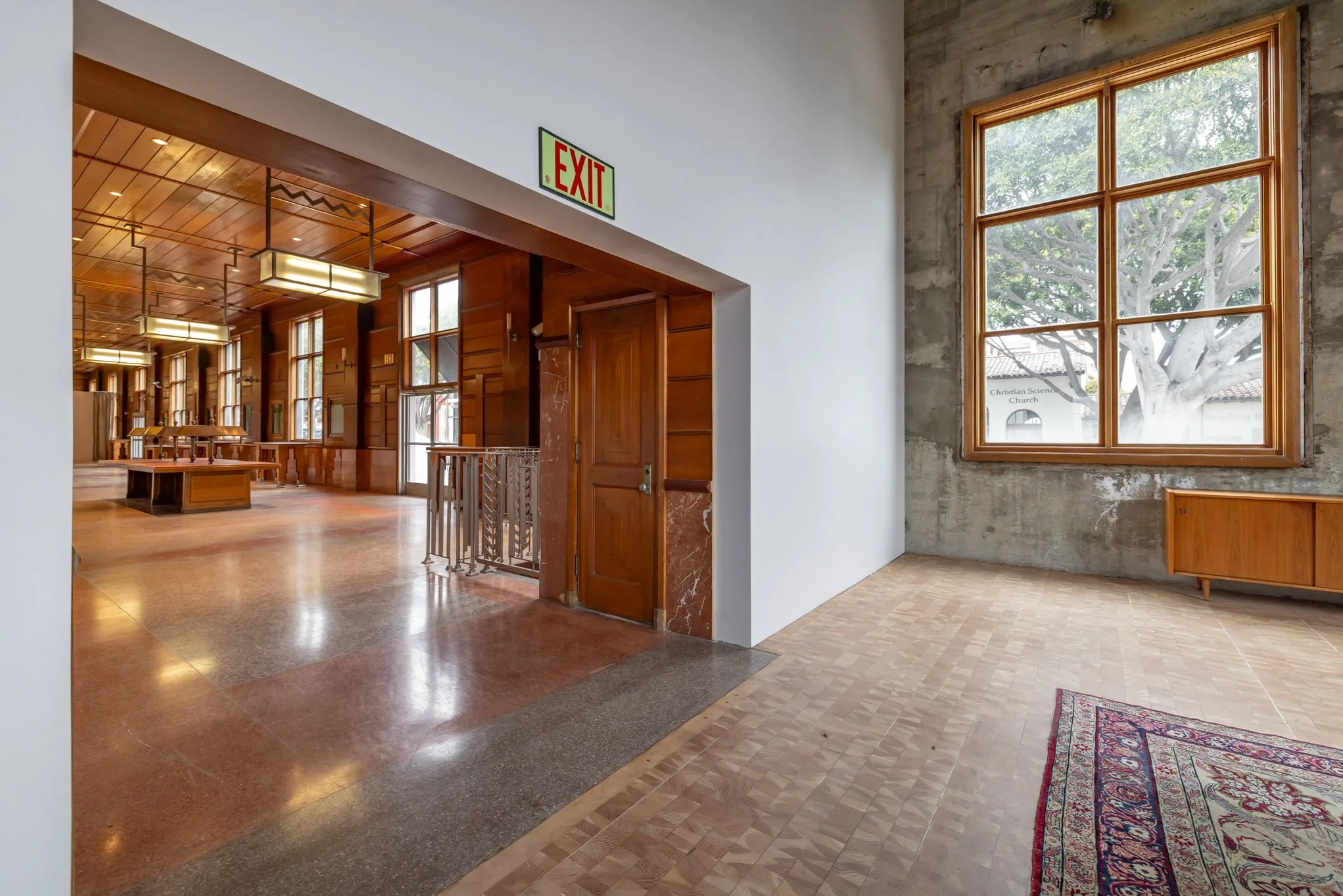 Interior of a building with a wooden ceiling, large windows, and a concrete wall, featuring an exit sign and a glimpse into a room with wooden furniture.