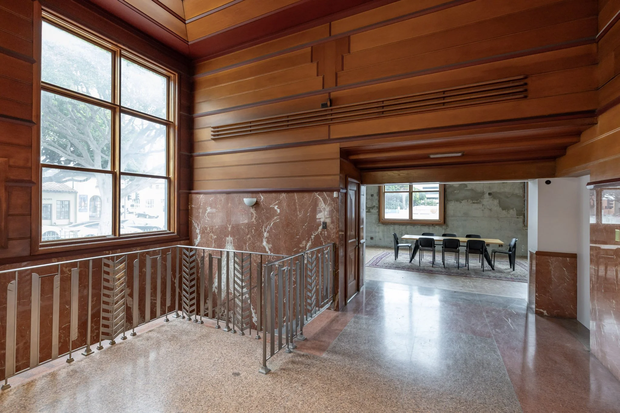 Interior of a building with wood-paneled walls, marble accents, large windows, a staircase with metal railing, and a meeting room with a long table and chairs.
