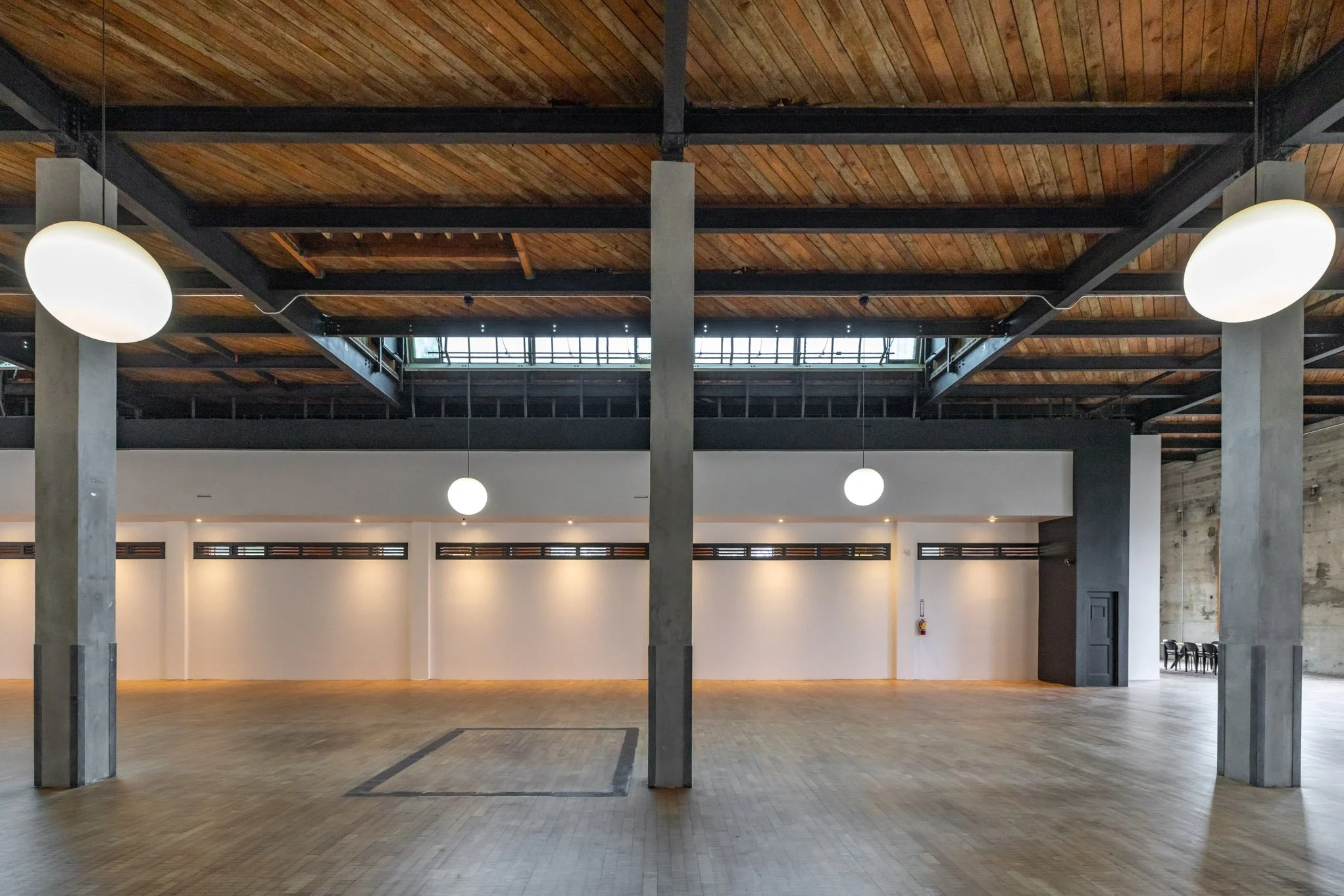 Empty spacious hall with wooden floor, white walls, exposed wooden ceiling, and hanging round light fixtures.