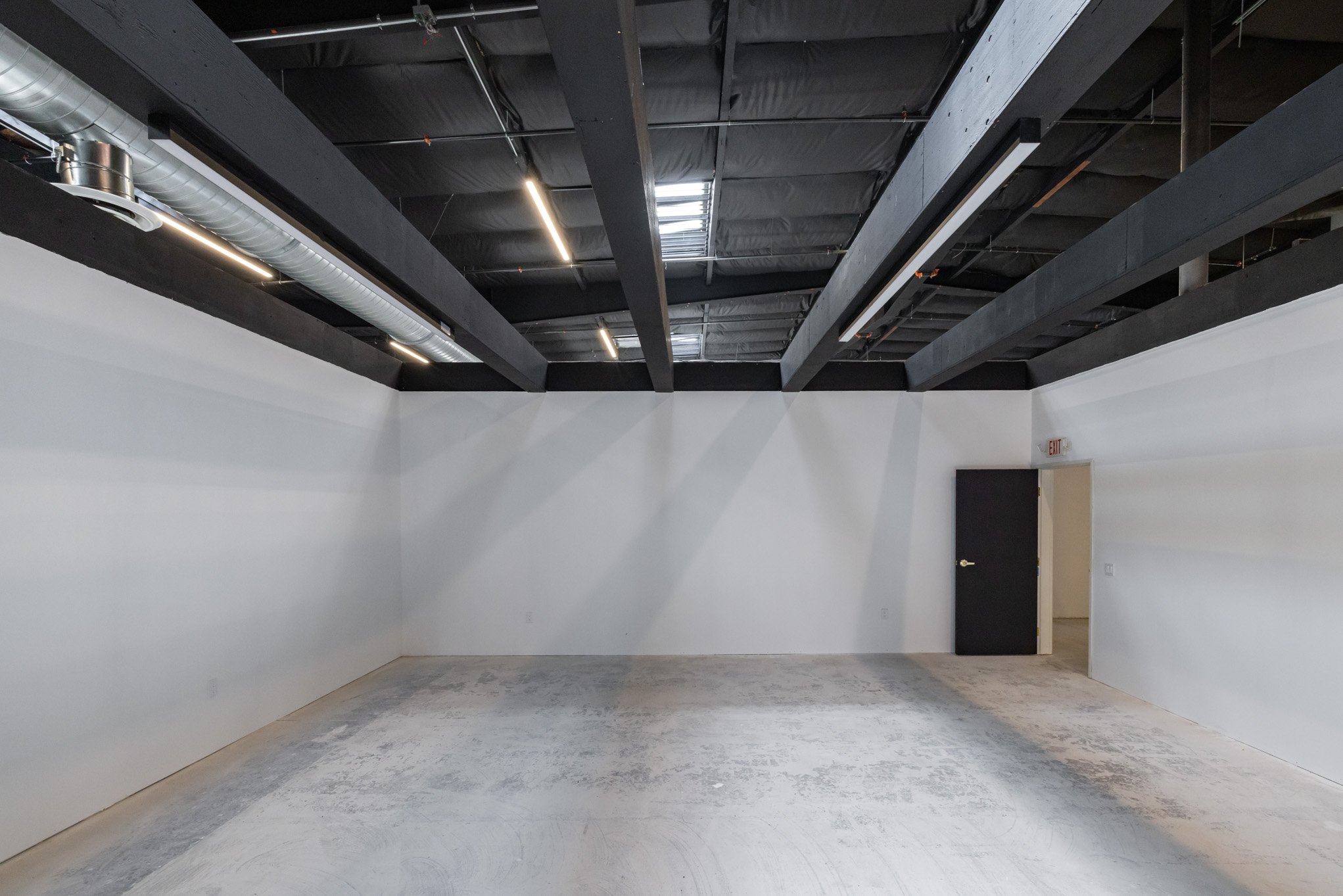 Empty room with white walls, concrete floor, exposed black ceiling beams, ductwork, and lighting fixtures, with an open door leading to another space.