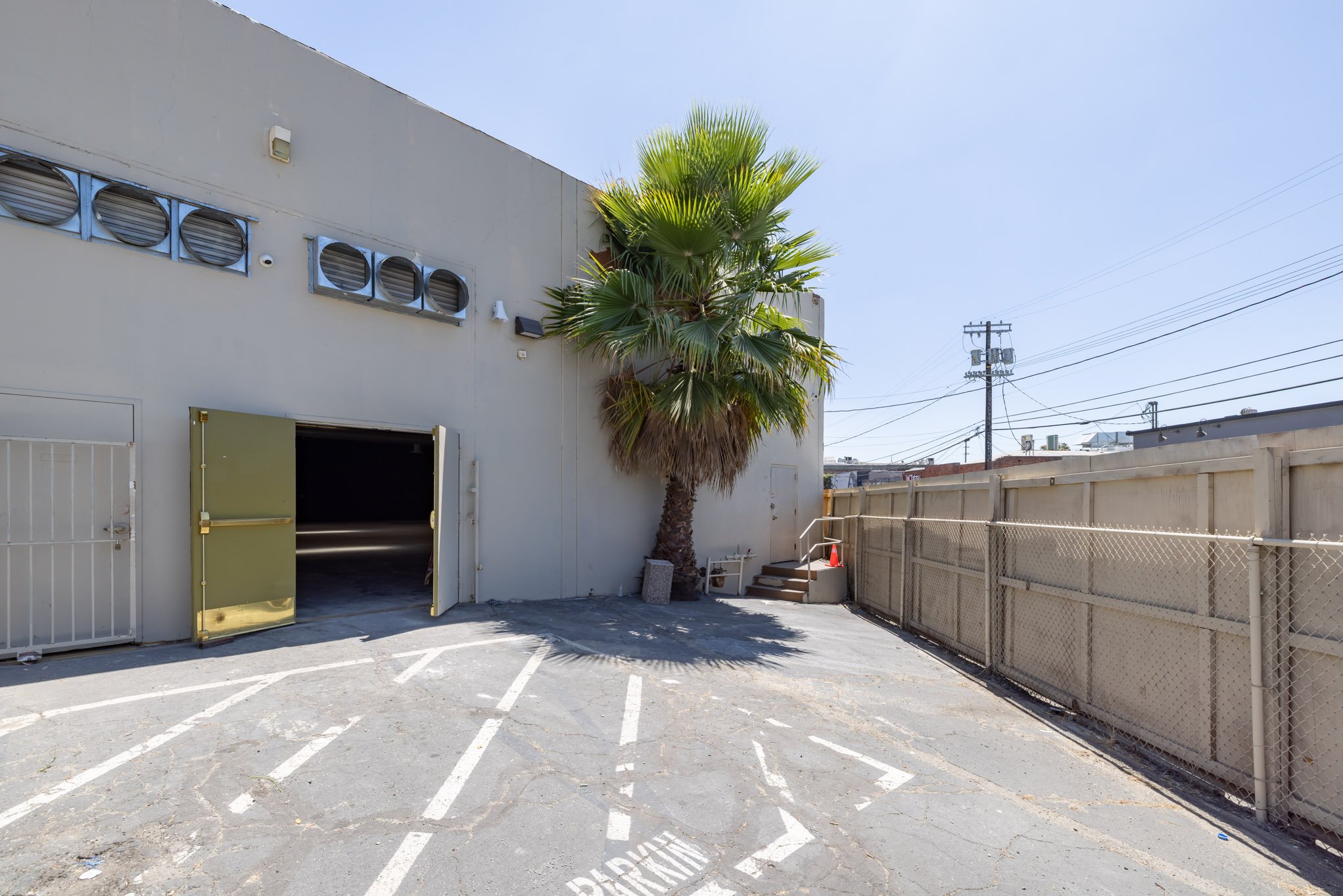 Empty parking lot with a palm tree near a beige building, open garage door, and power lines in the background.