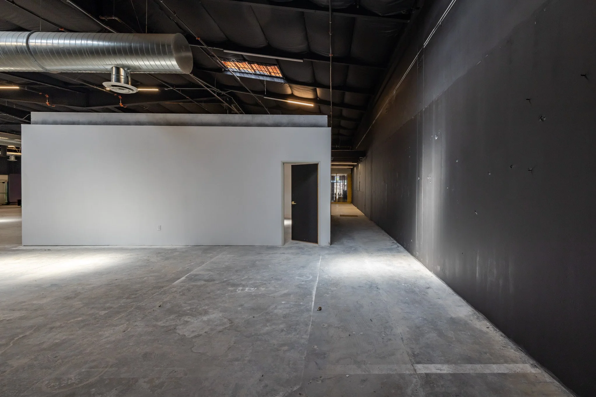 Empty industrial or commercial space with concrete floor, black wall on the right, white wall with doorway in the center, exposed ductwork and ceiling pipes overhead.