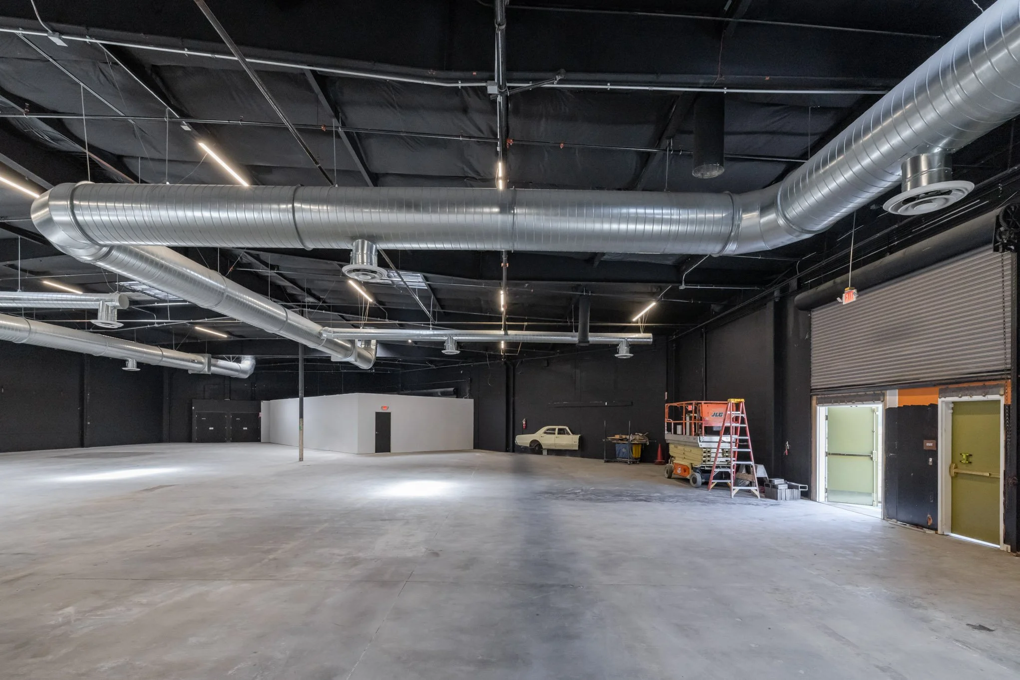 An empty industrial commercial space with black walls, silver ductwork on the ceiling, two open metal garage doors, a small white vintage car, and maintenance equipment including a lift, ladder, and carts.