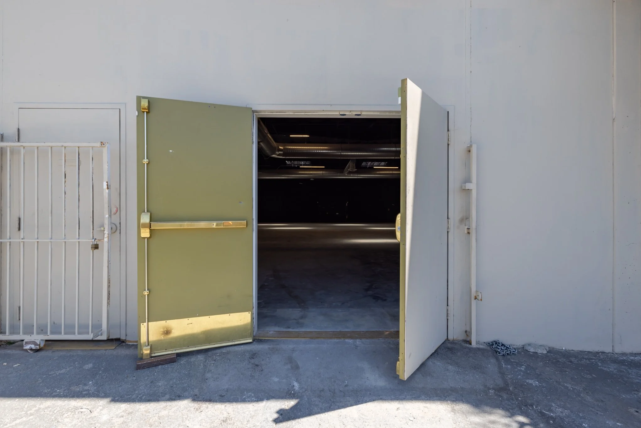 Open garage door revealing an empty storage or service area with metallic pipes on the ceiling, along a concrete floor, outside a beige building with a white fence on the left.