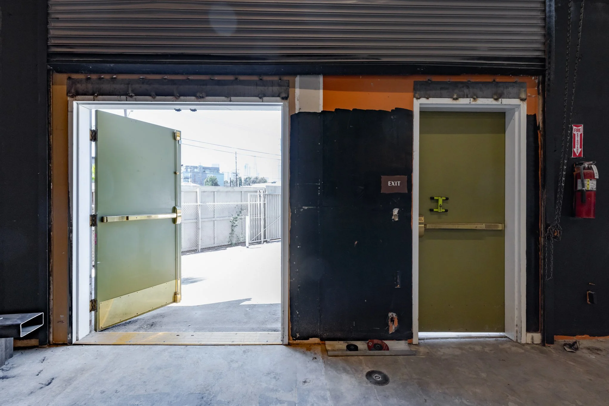 A partially finished interior with an open garage door on the left, leading outside, and a small door marked "EXIT" on the right, revealing a bright outdoor area with a fence and some buildings in the distance. There is a fire extinguisher mounted on