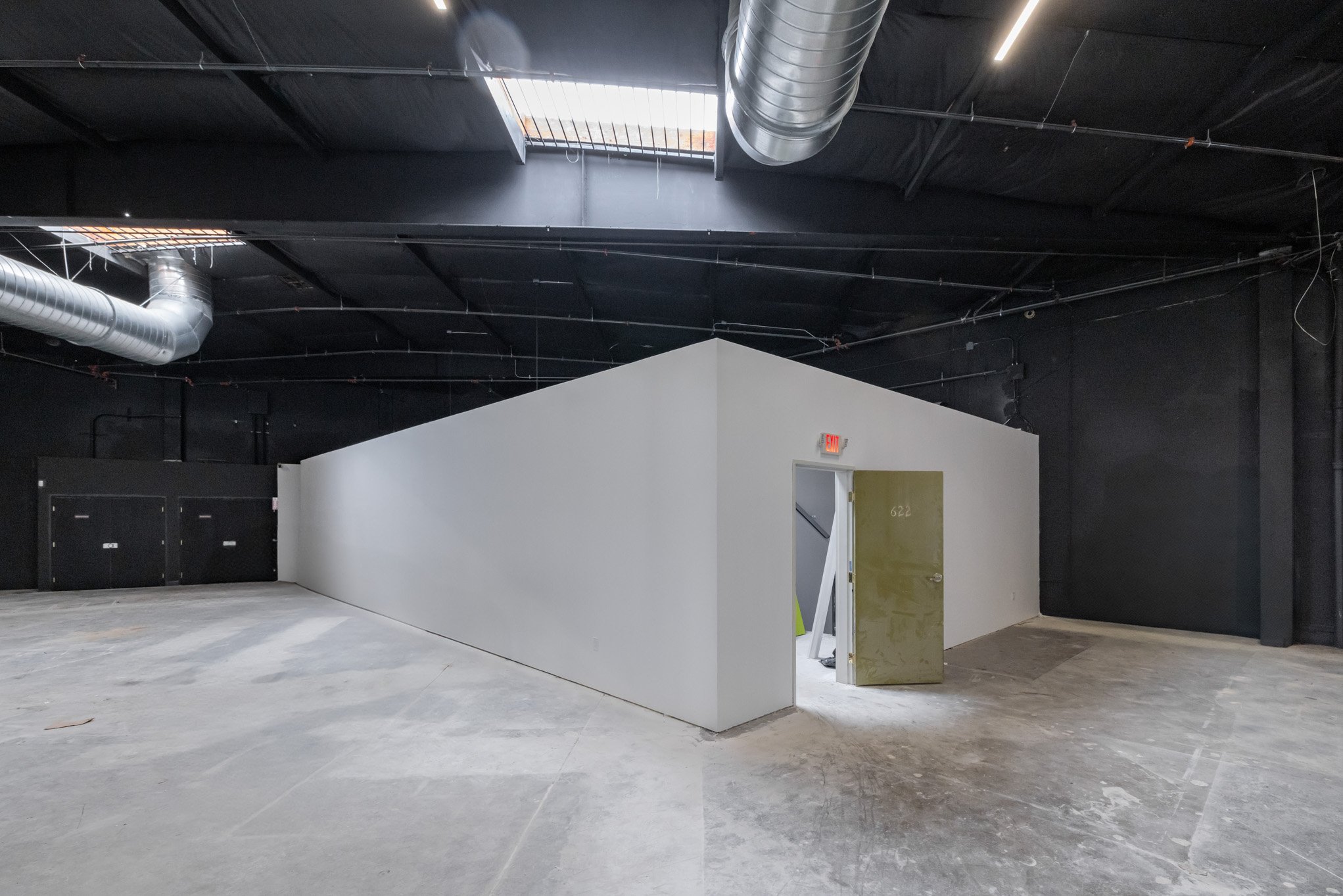 Interior view of a construction site with a small white partition wall, a partially open door with the number 622, black and metallic ductwork on the ceiling, and an open skylight.
