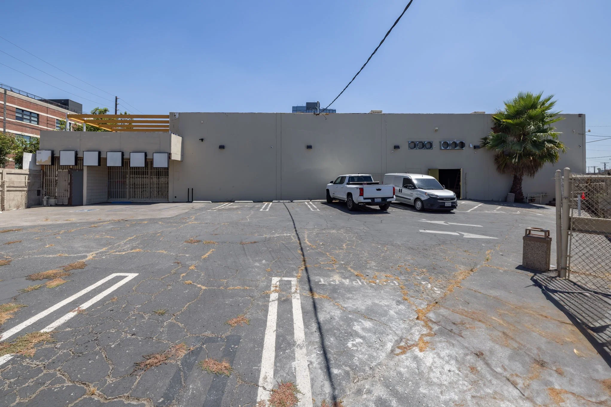 Empty parking lot with two white trucks and a gray van parked in front of a beige building with a palm tree on the right side.