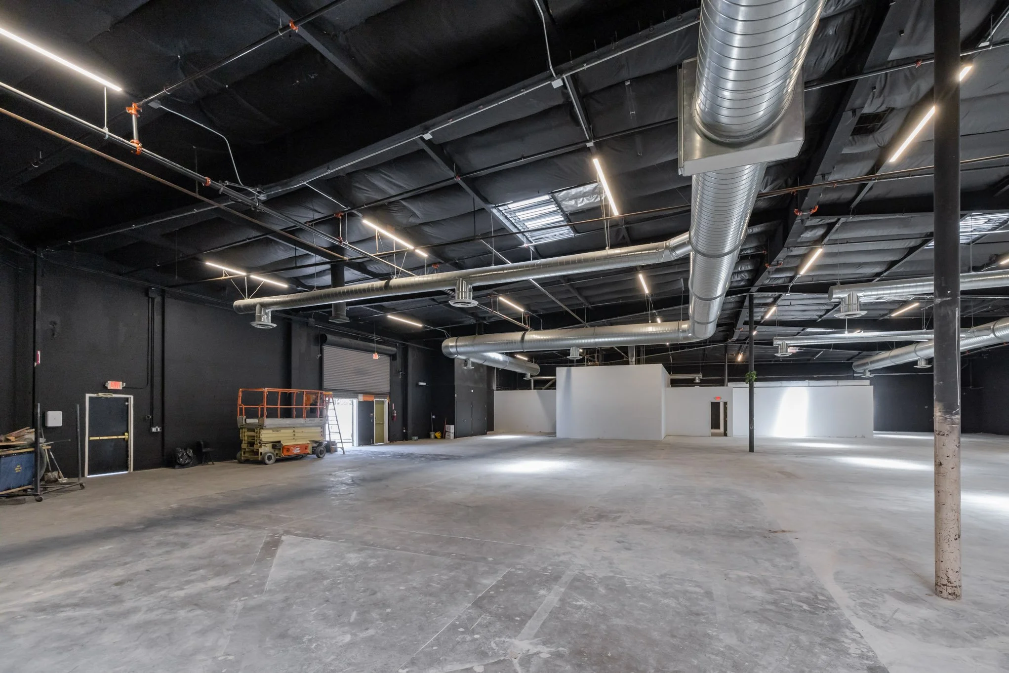 Large empty warehouse with black walls, exposed ductwork, and construction equipment near the back wall.