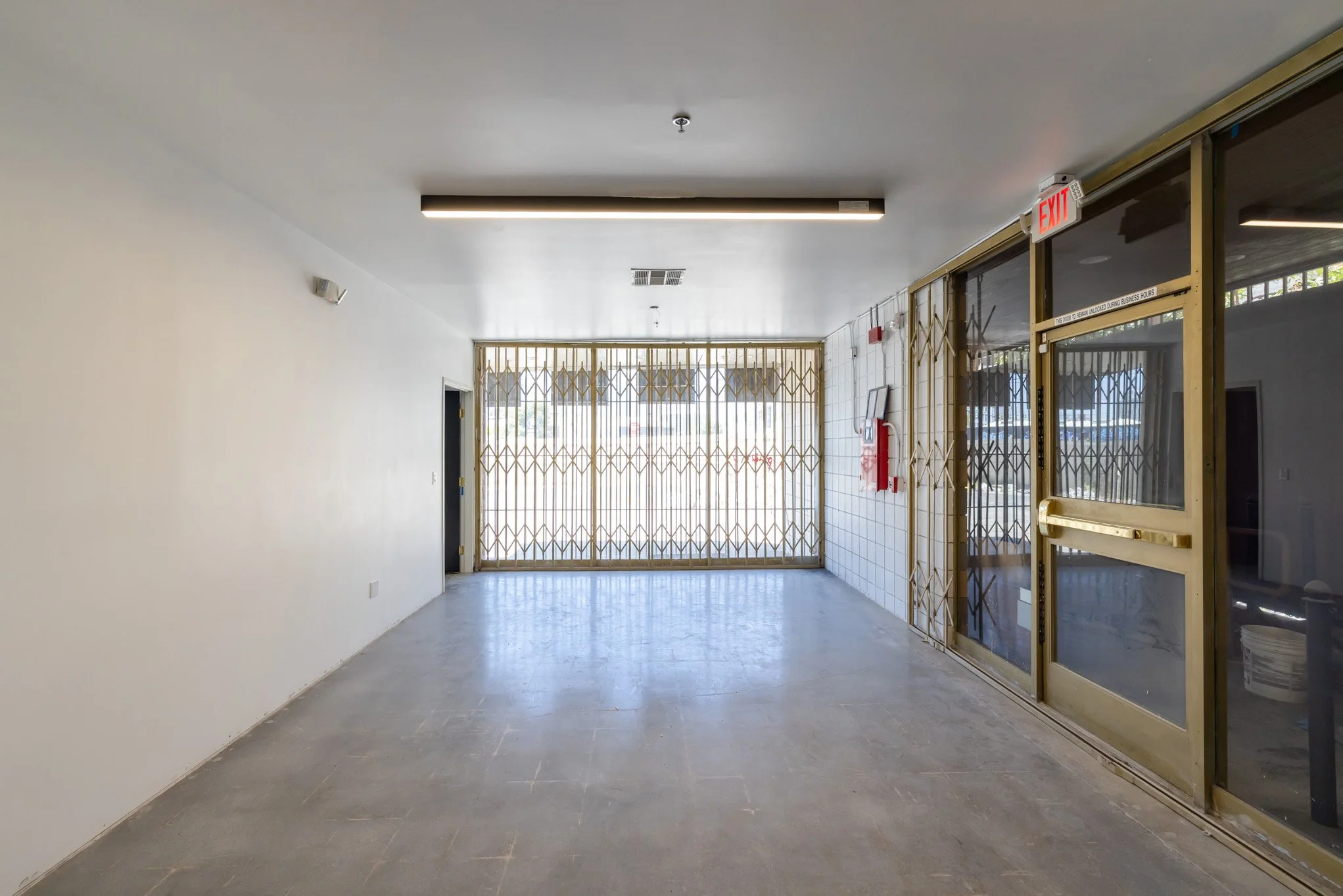 Empty indoor corridor with a closed metal gate at the end, glass doors on the right, and a white wall on the left.