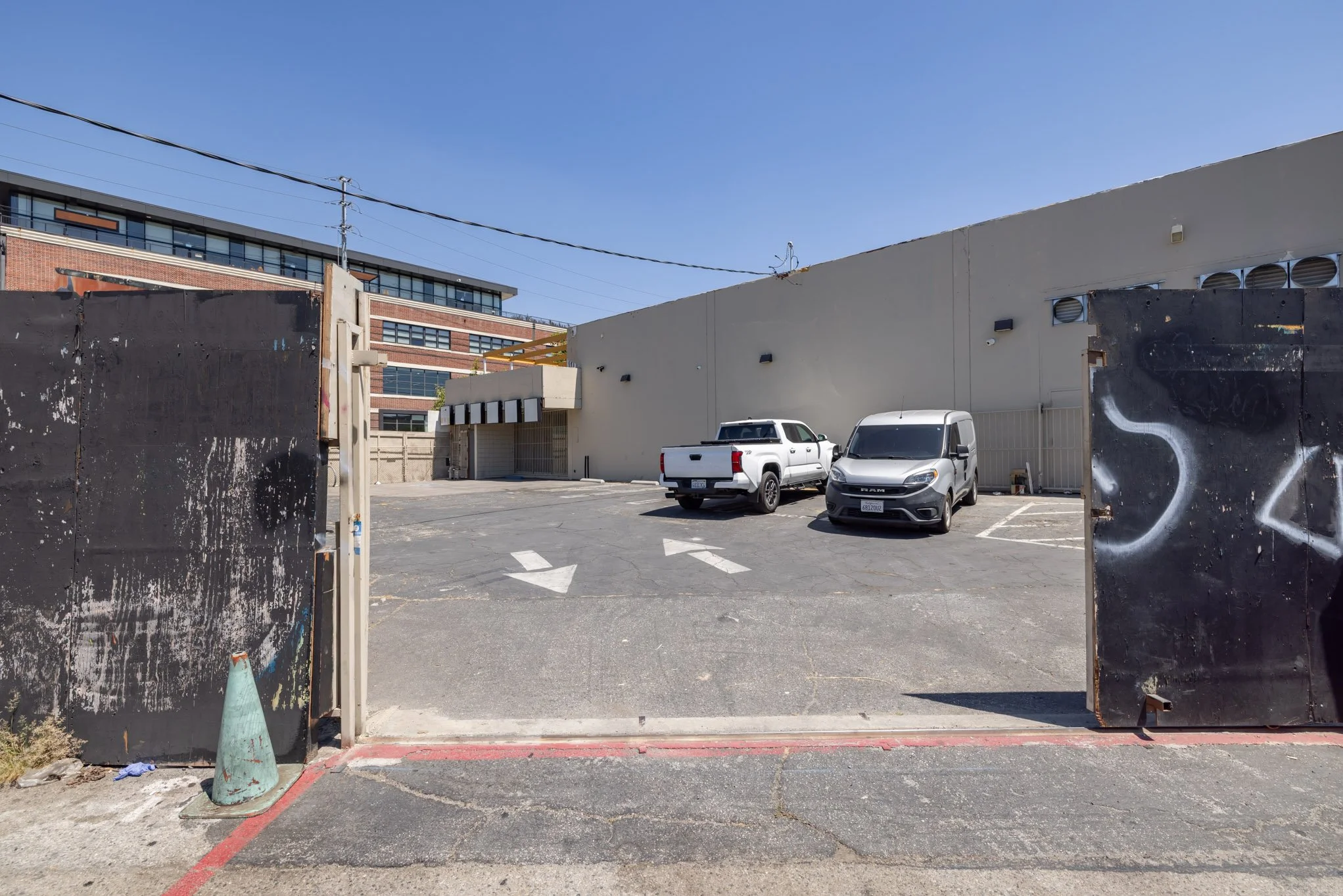 A parking lot viewed from behind a black metal gate with graffiti, with two parked trucks and a building in the background under a clear blue sky.