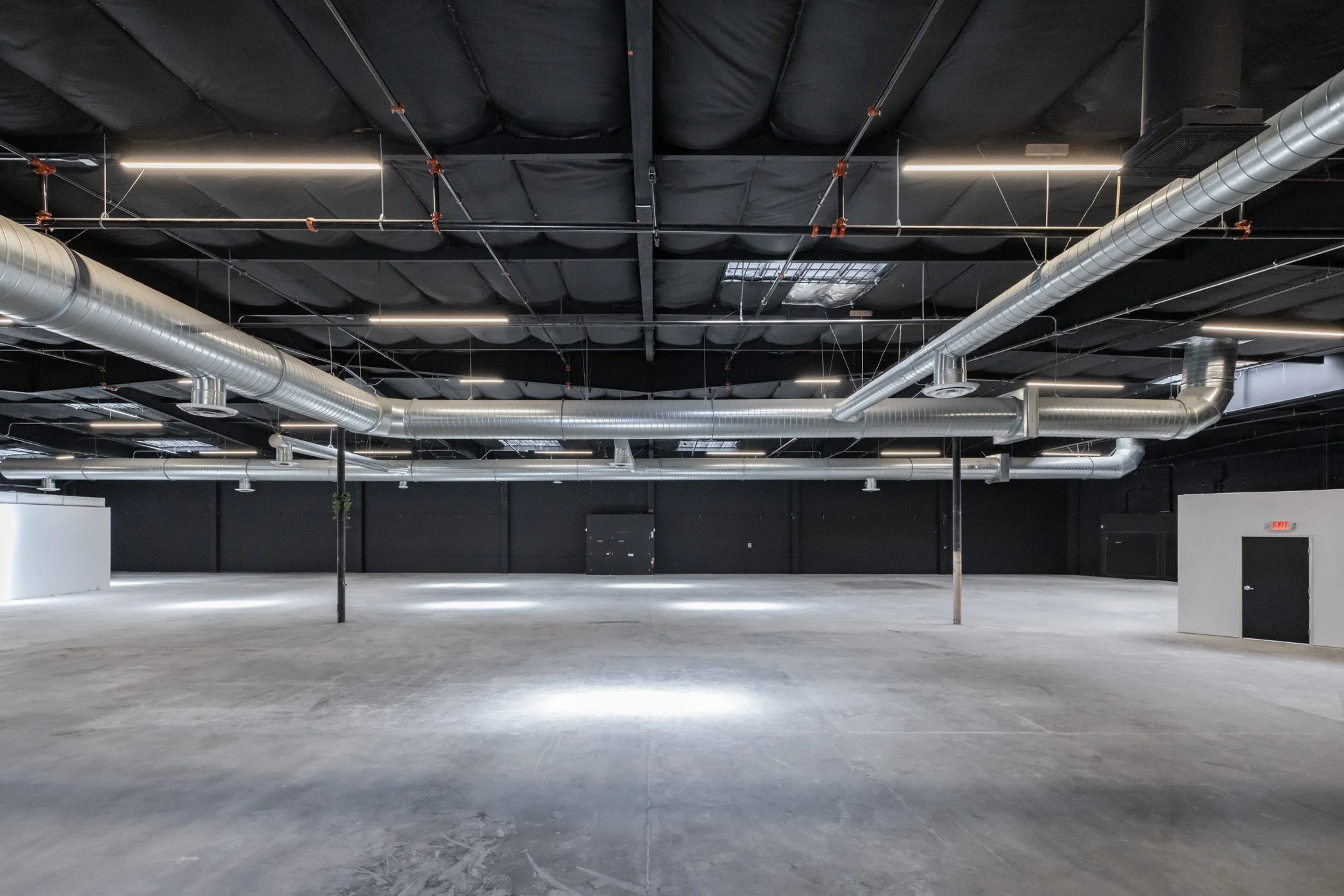 Empty commercial or industrial space with a concrete floor, black ceiling with exposed ducts and pipes, and a few small objects hanging from the ceiling.