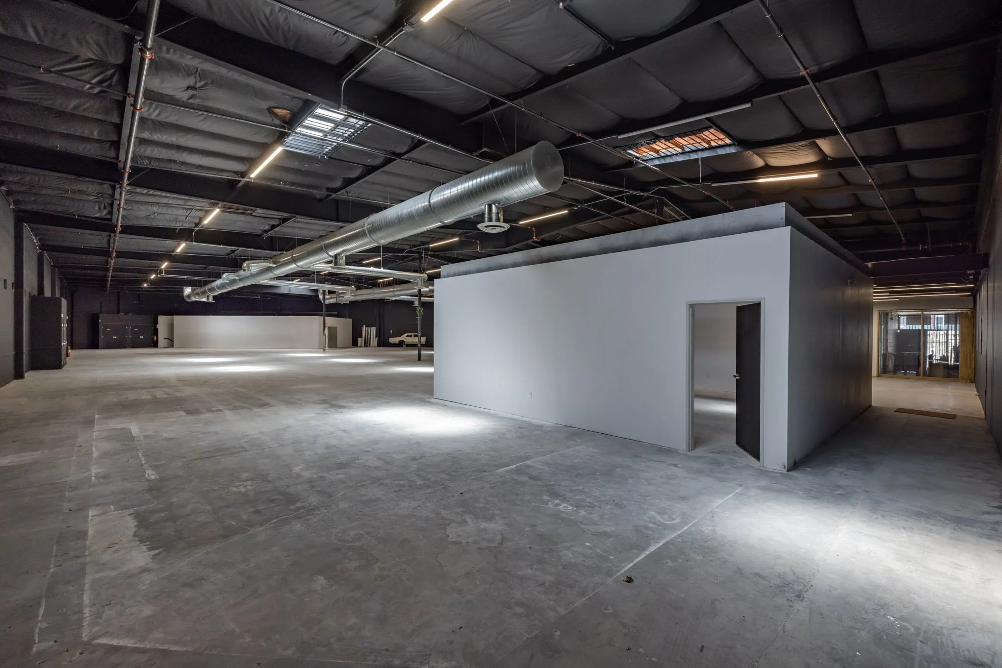 Empty commercial interior with a small white room, unfinished concrete floor, and black ceiling with exposed ductwork and lighting.