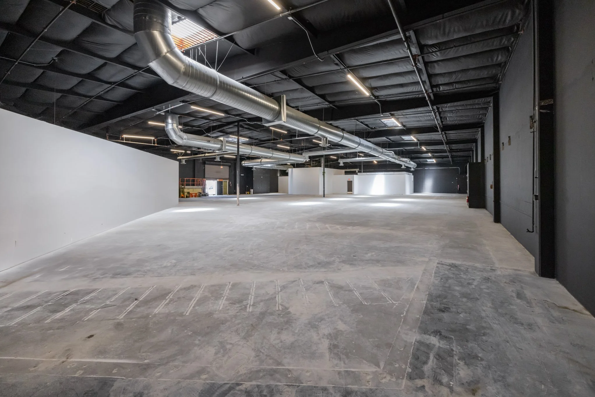 Empty, large commercial space with unfinished concrete floors, black ceiling with exposed ductwork, and black and white walls. Construction equipment and doors are visible in the background.