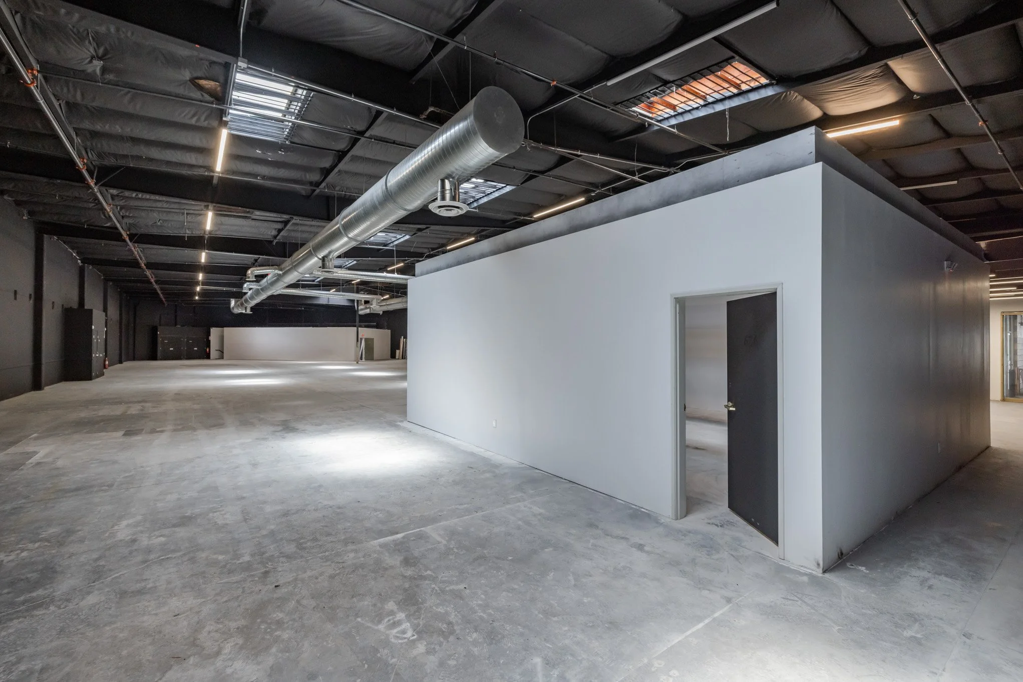Empty commercial space with unfinished concrete floors, black ceiling with exposed ductwork, and a small enclosed room with a white wall and an open door.