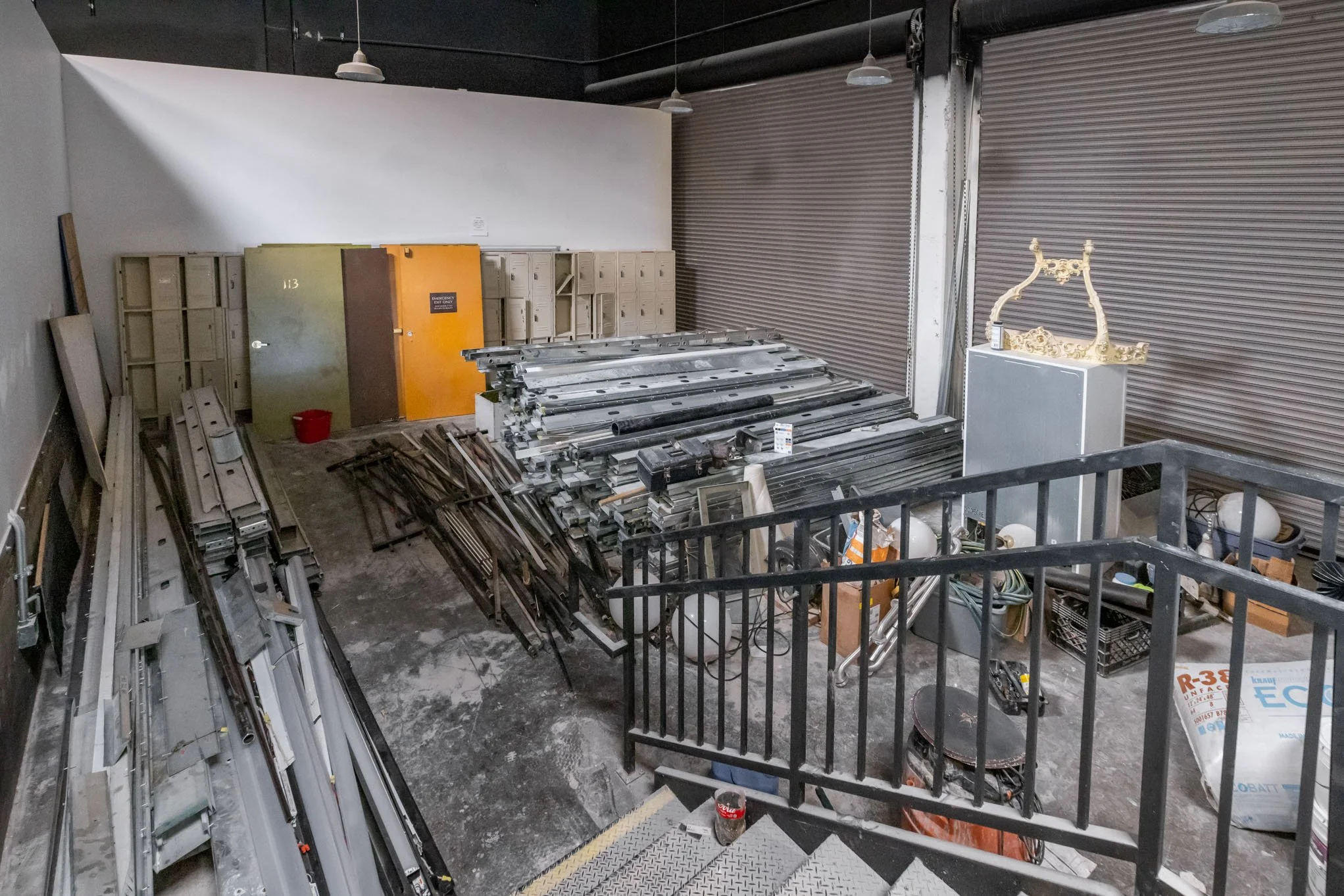 Storage room with metal pipes, electrical components, lockers, and decorative items, including a gold-colored ornate frame or crown, behind a railing in an industrial setting.