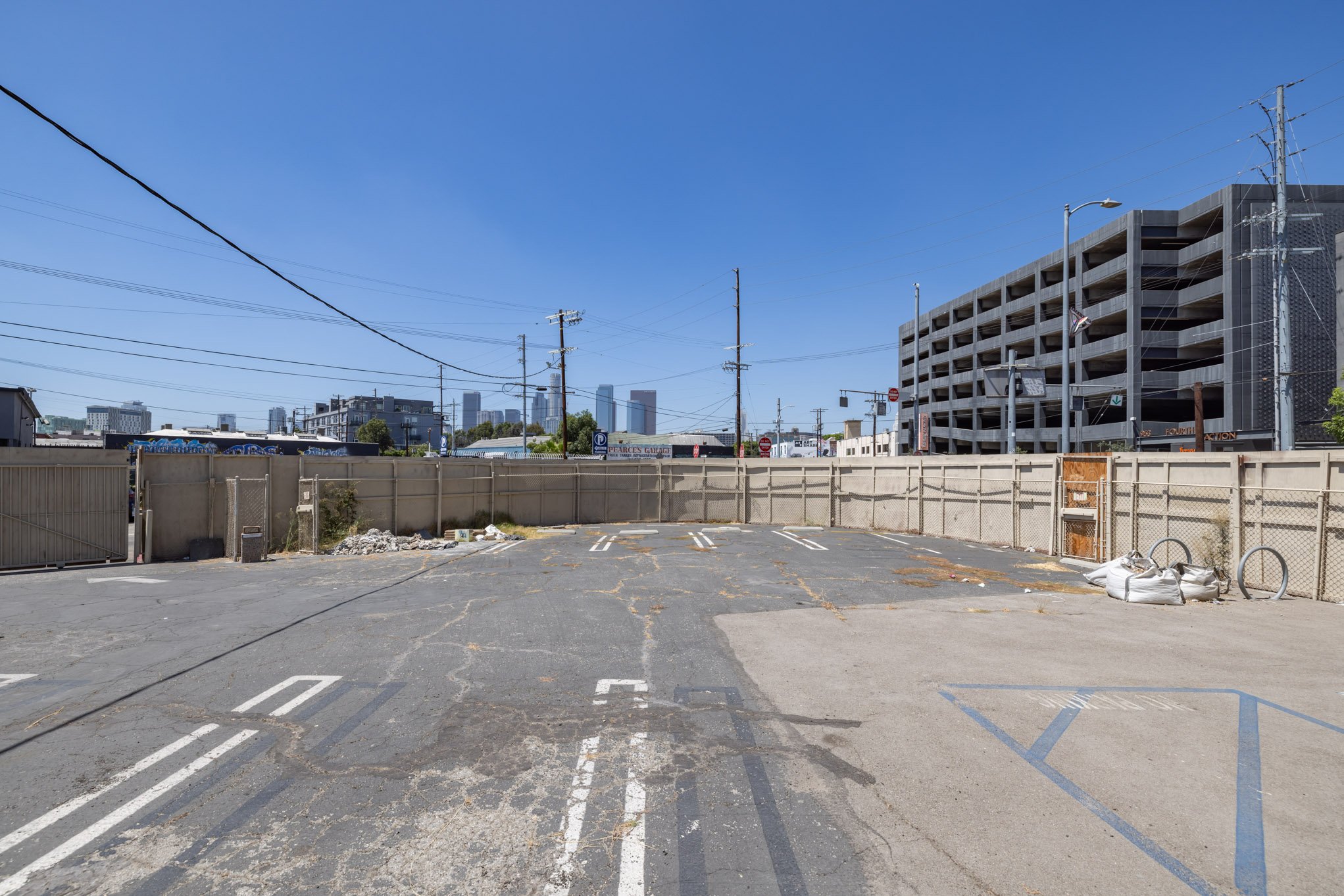 An empty parking lot with cracked asphalt and old white painted parking lines, surrounded by chain-link fencing, with a city skyline and office building under a clear blue sky in the background.