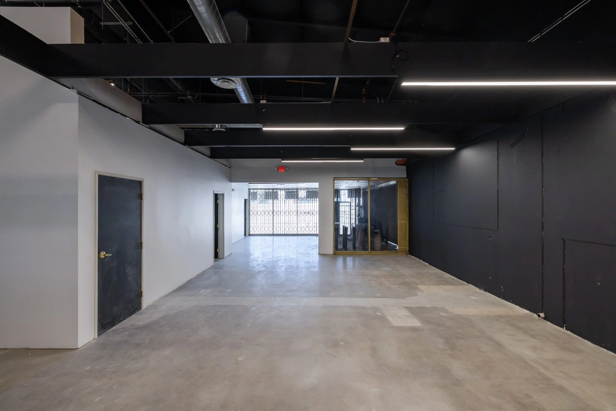 Empty commercial space with black and white walls, exposed ceiling pipes, and concrete flooring. Front entrance has metal security gates and glass door with gold trim.