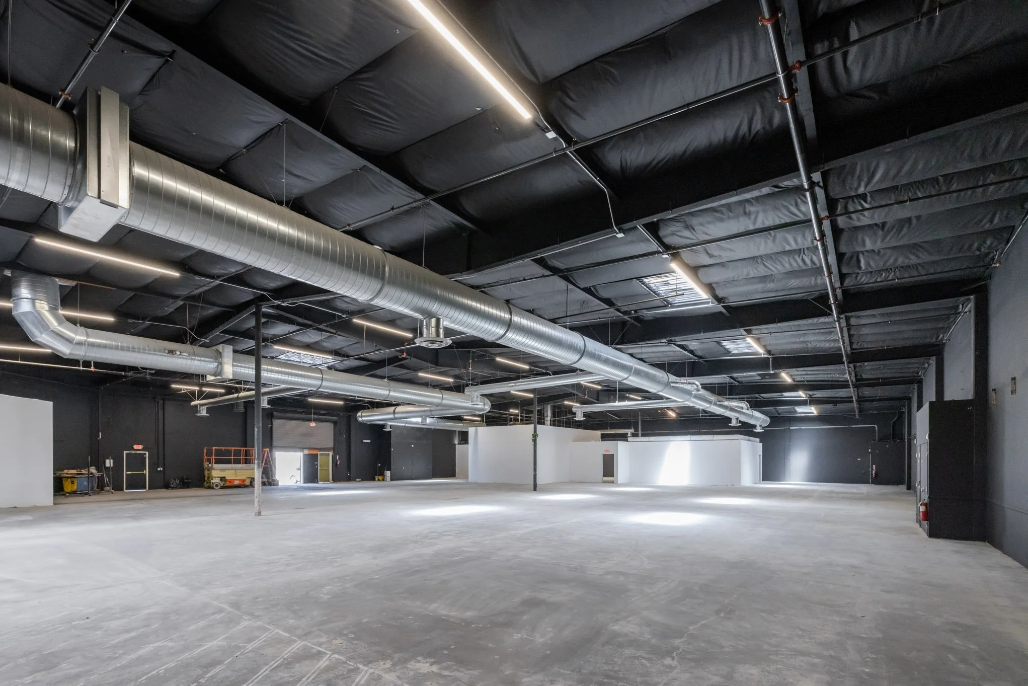 Empty industrial warehouse with black ceiling insulation, exposed gray metal ducts, and concrete floor.