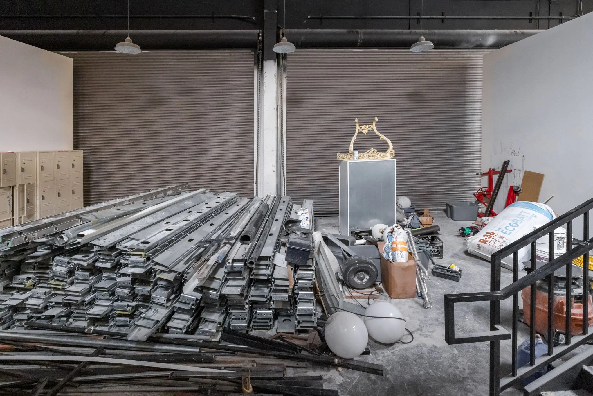 Storage area with construction materials, including a large pile of metal frames, scattered light fixtures, boxes, and tools, in front of a closed industrial metal shutter.