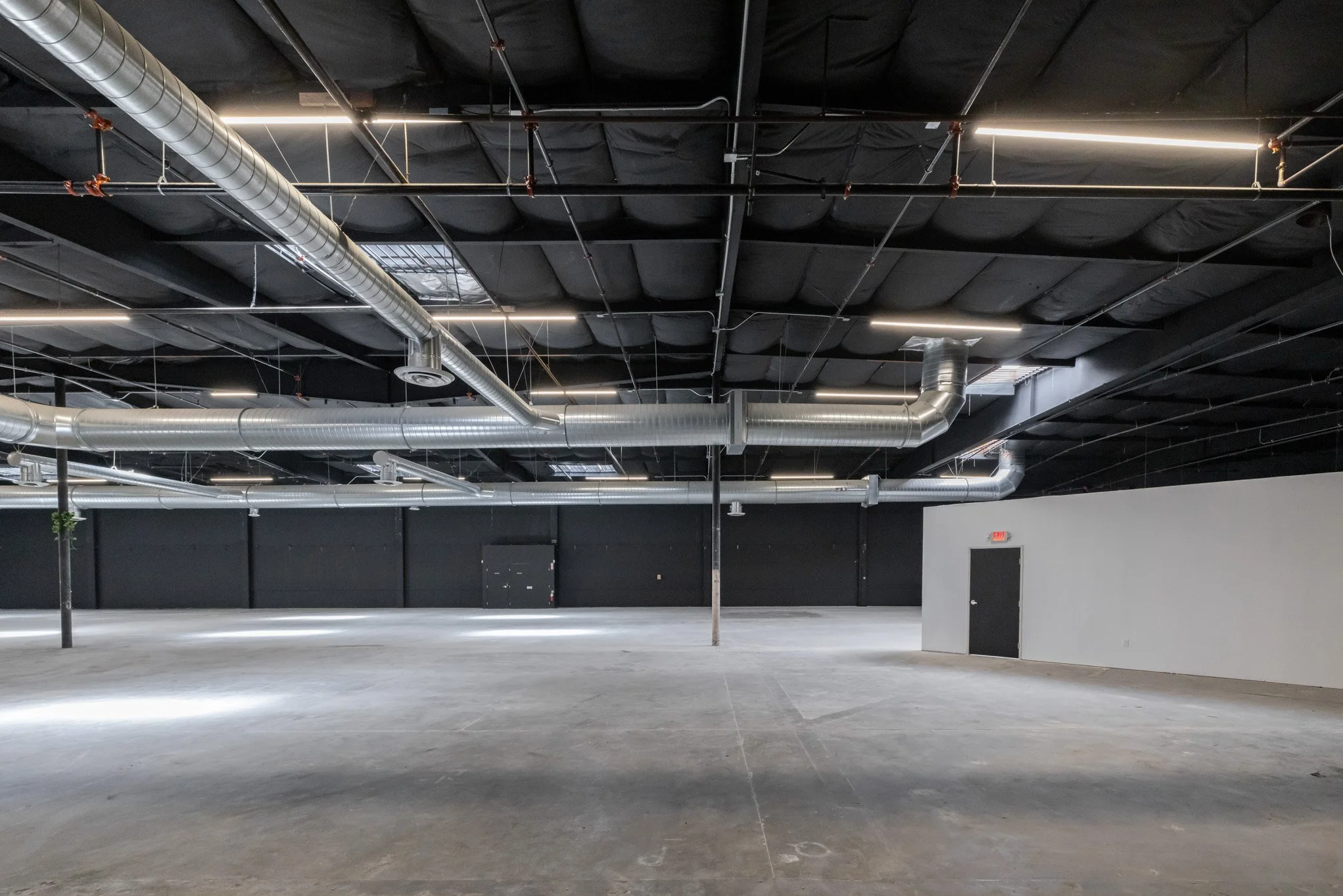 Empty industrial or commercial warehouse space with exposed ceiling pipes, ductwork, and LED lighting, featuring a concrete floor and black and white walls.
