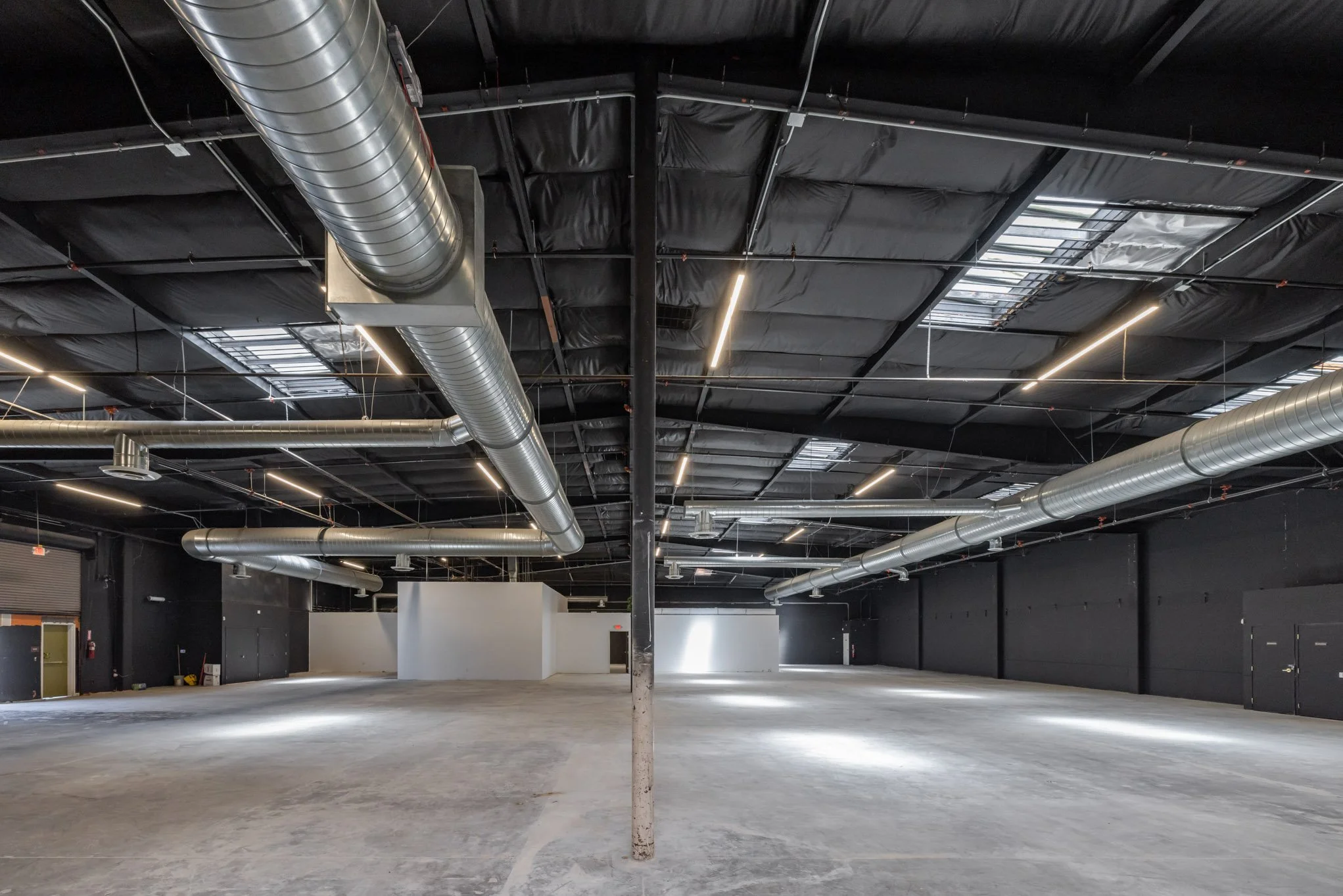 Empty indoor space with black walls, exposed ductwork, large industrial ceiling, and minimal lighting.