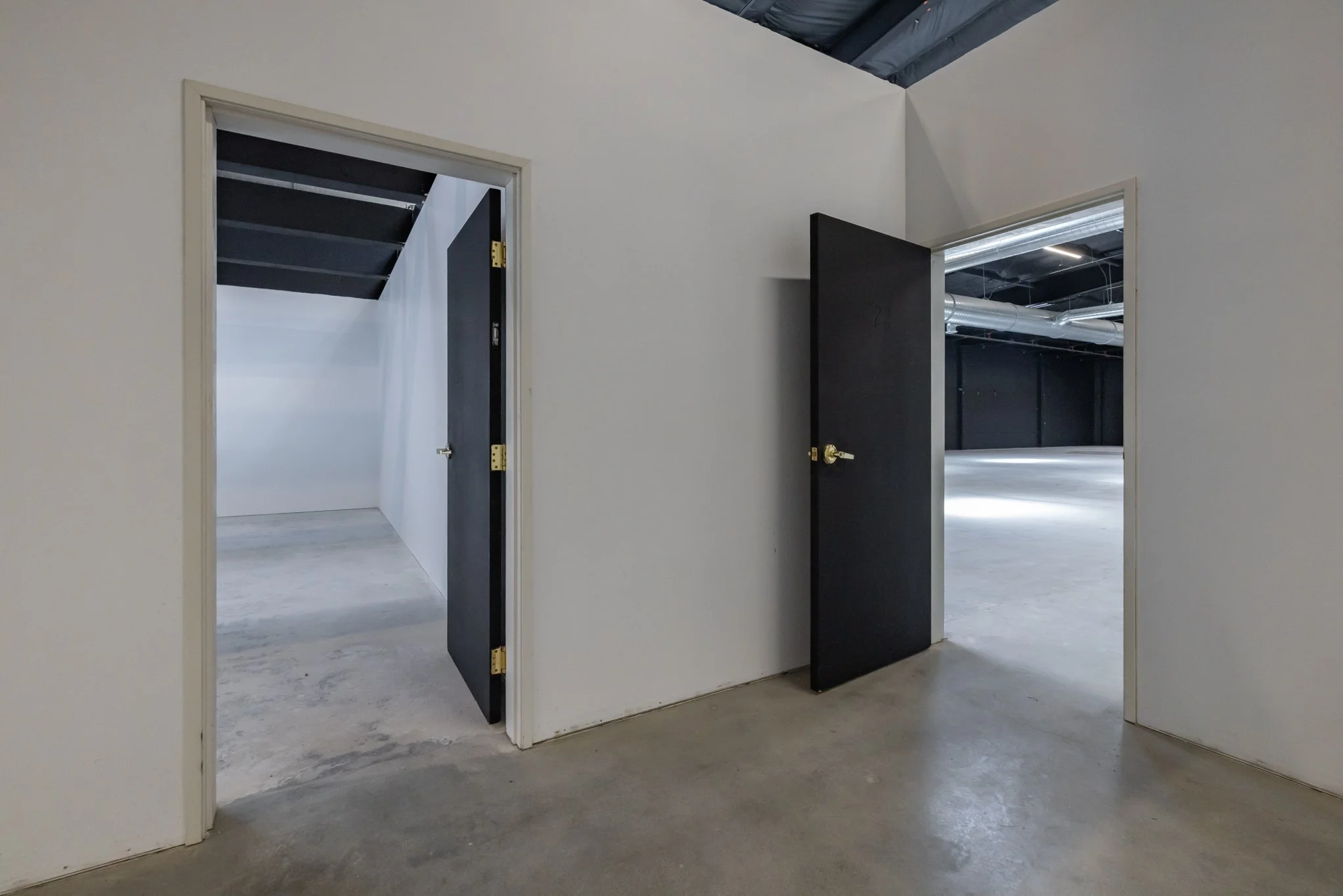Interior of a building with two open doors leading to empty storage spaces or rooms, with concrete floors and black ceiling pipes visible through the doors.