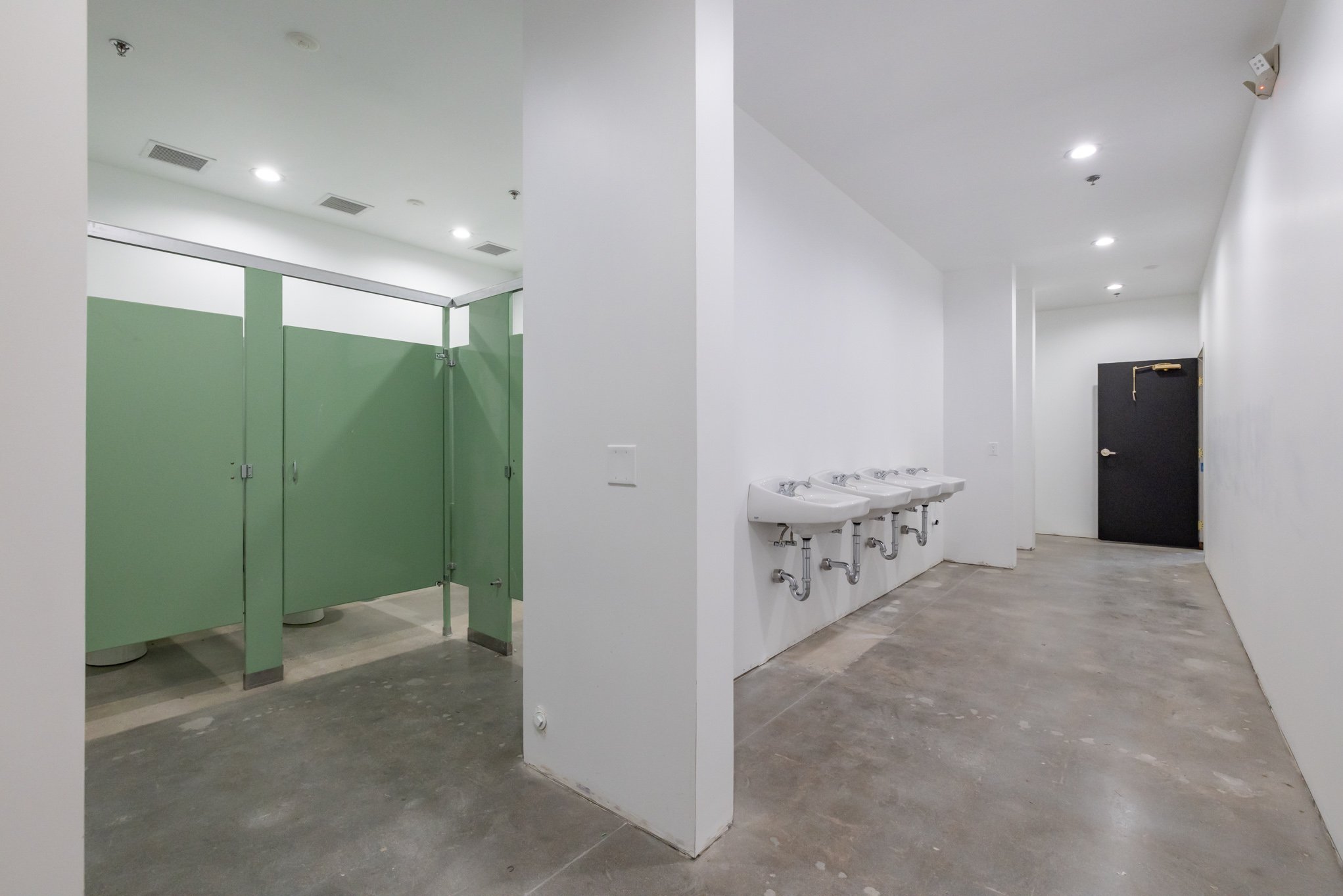 A public restroom with green stalls on the left and a row of sinks on the right. The room has a concrete floor, white walls, and ceiling, with overhead lighting and a black door at the far end.