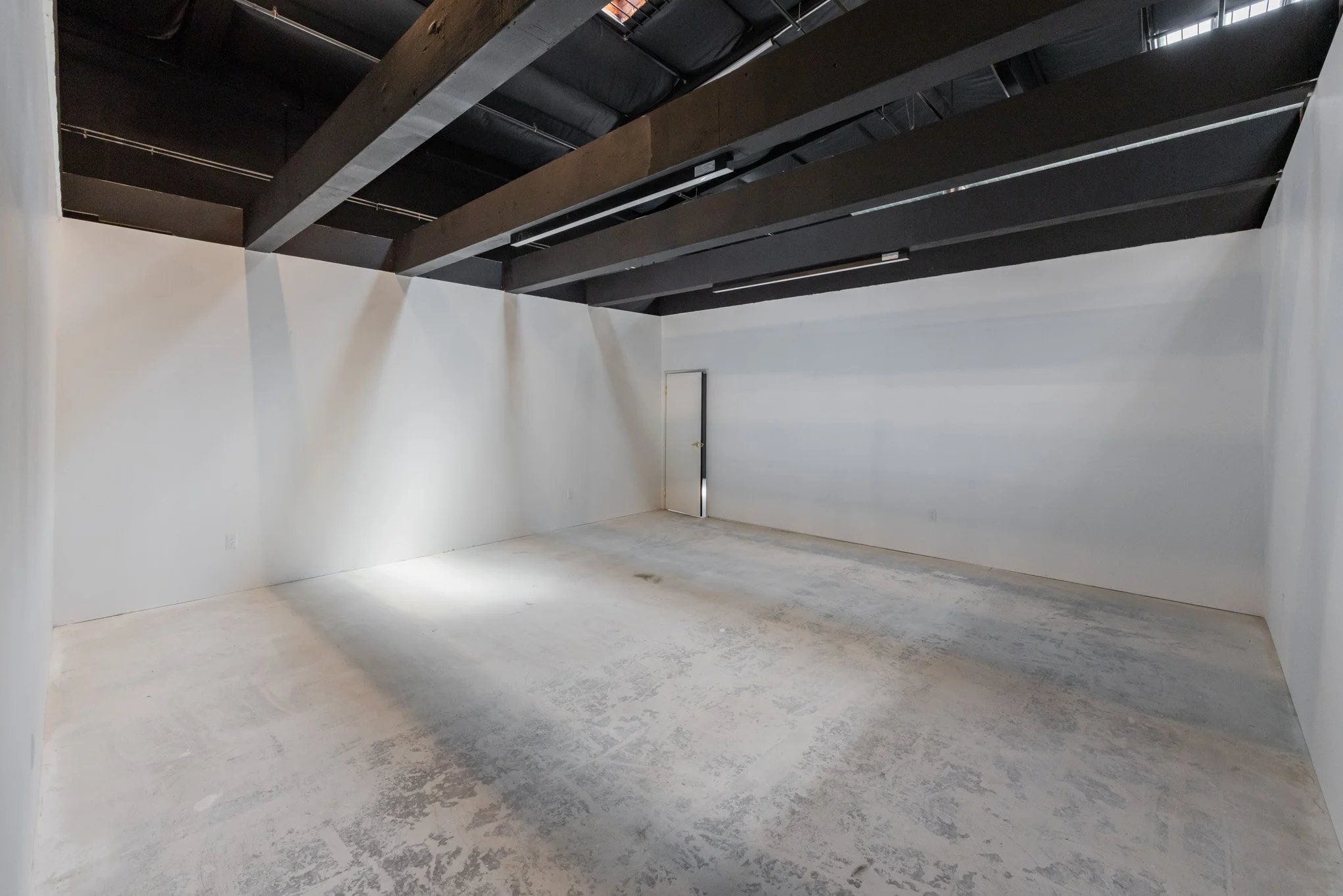 Empty white-walled room with an industrial ceiling and a door, possibly in a warehouse or studio space.