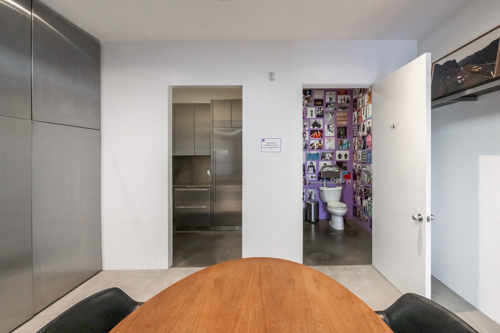 A modern interior room with a wooden table and black chairs, with a doorway leading to a bathroom with purple walls decorated with photos, a toilet, a metal trash can, and a small wastebasket.