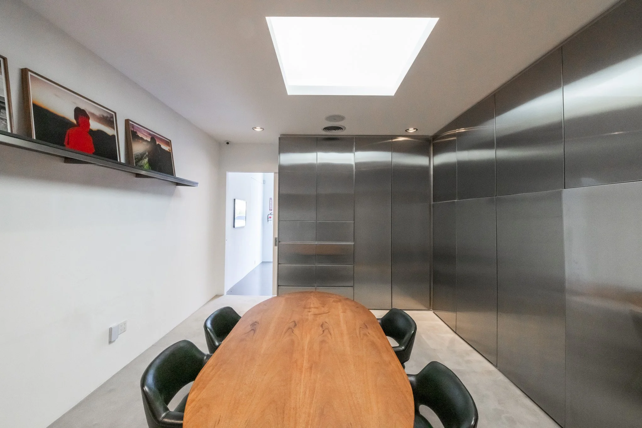 Modern conference room with a wooden table, black chairs, metallic wall panels, and framed photographs on a shelf.