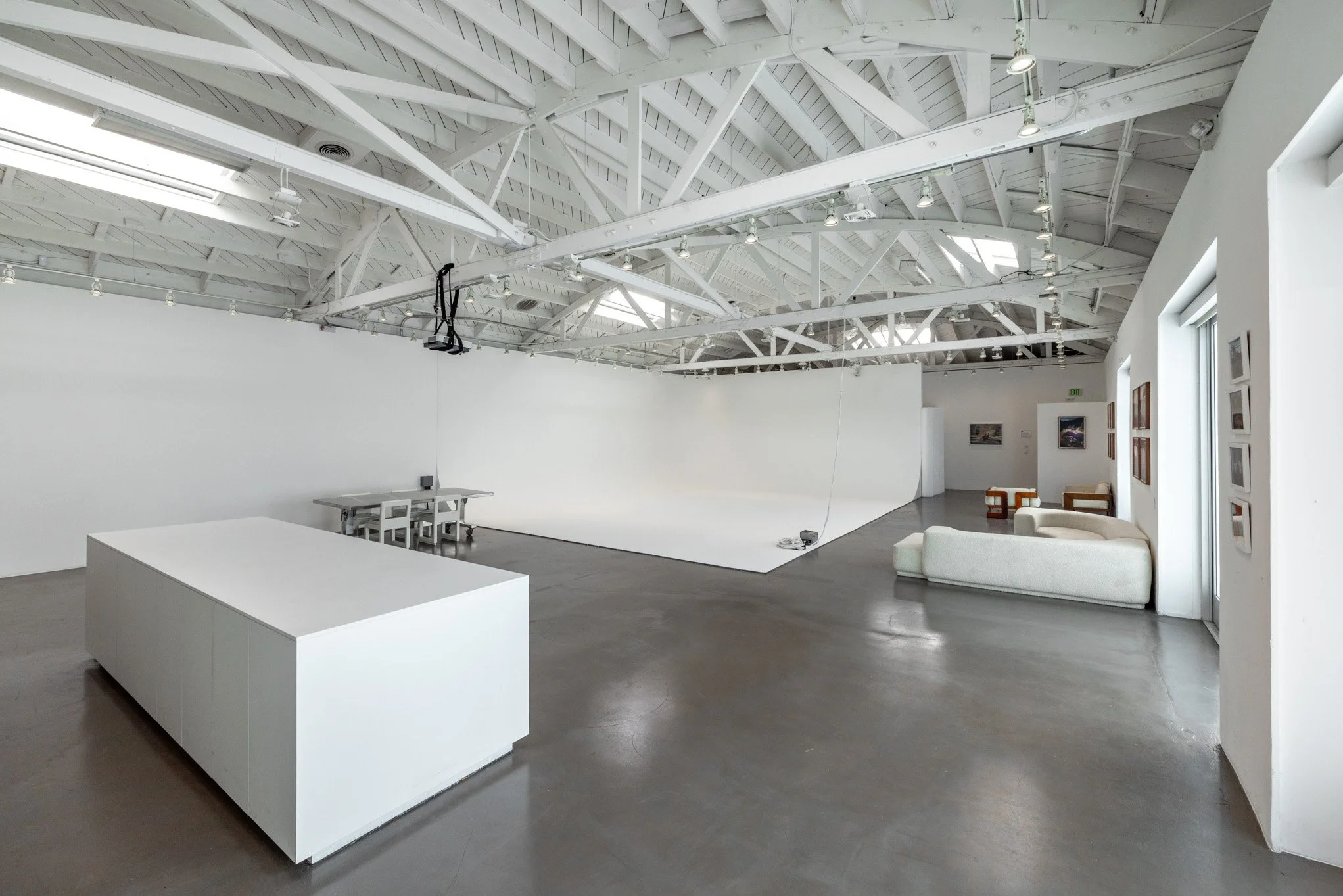 Empty art gallery with white walls, high ceiling with exposed beams, and a polished concrete floor. Contains white display furniture and framed pictures on the walls.