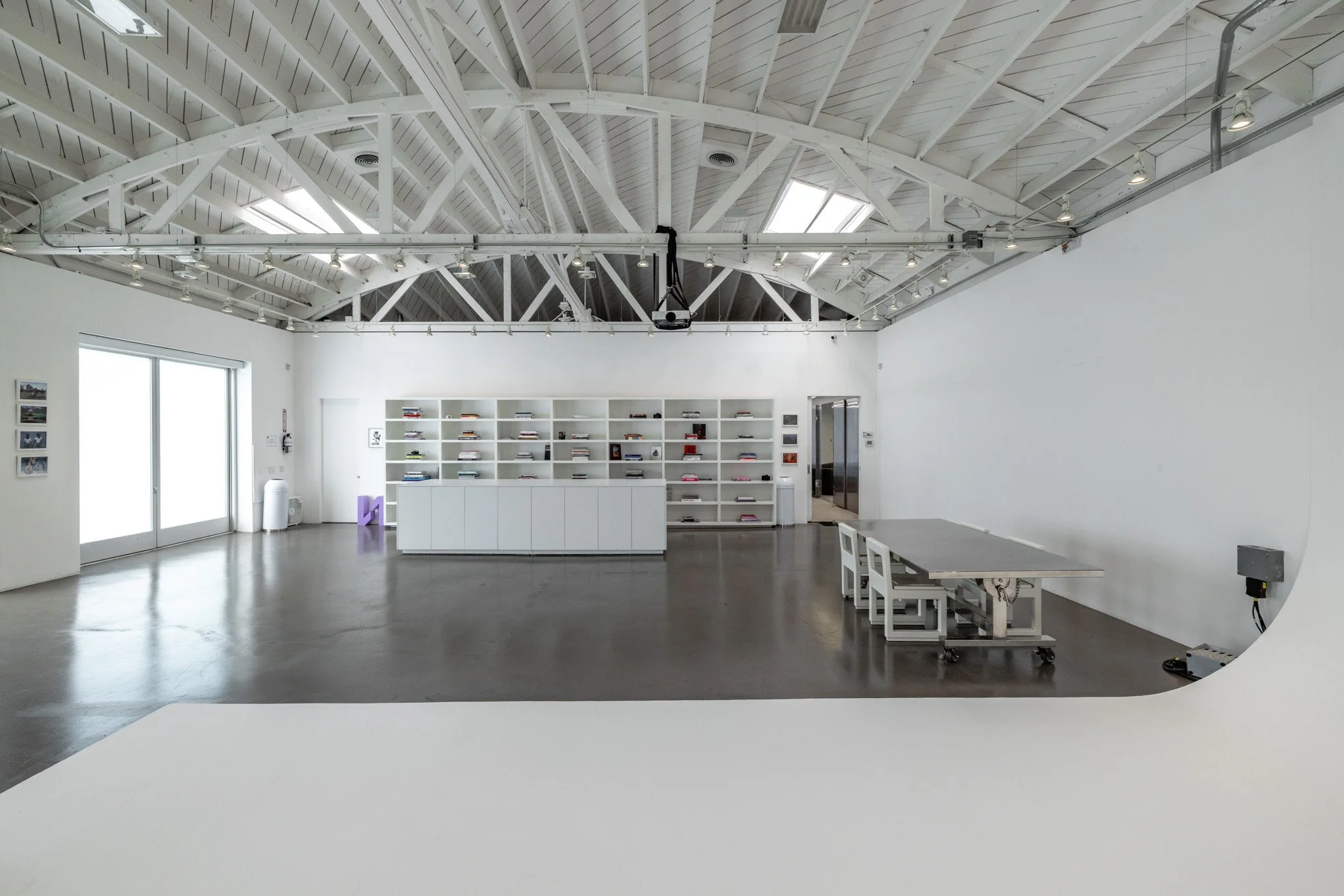 Empty art gallery with white walls, a high skylit ceiling, a large window, and a bookshelf filled with books in the back