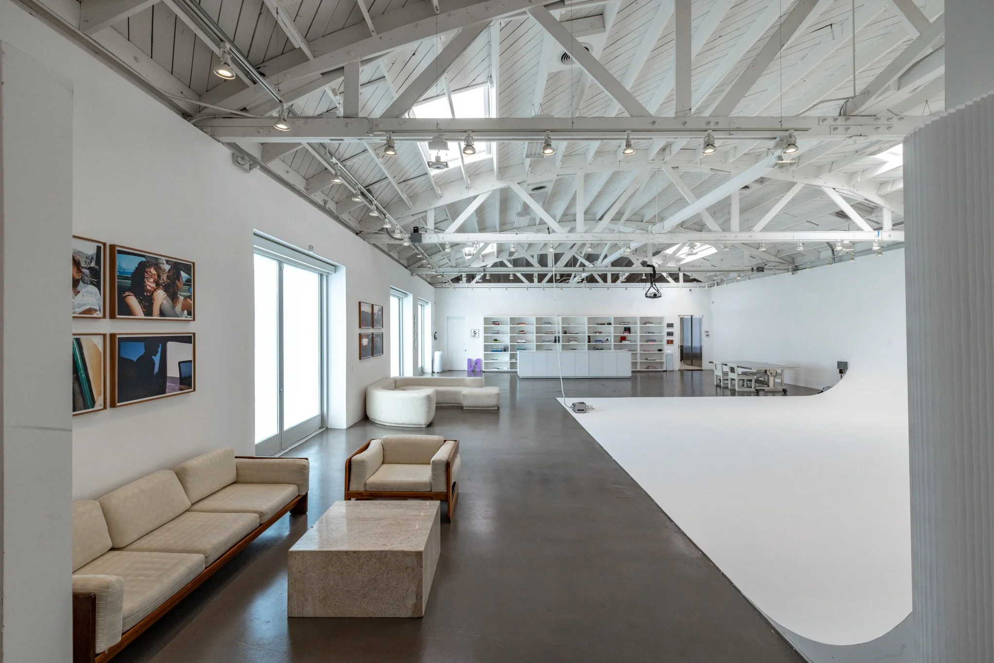 Spacious interior of a modern photo studio with white walls, exposed ceiling beams, and ample natural light. Features seating area with cream sofa and chairs, wall art, and a large white cyclorama wall.