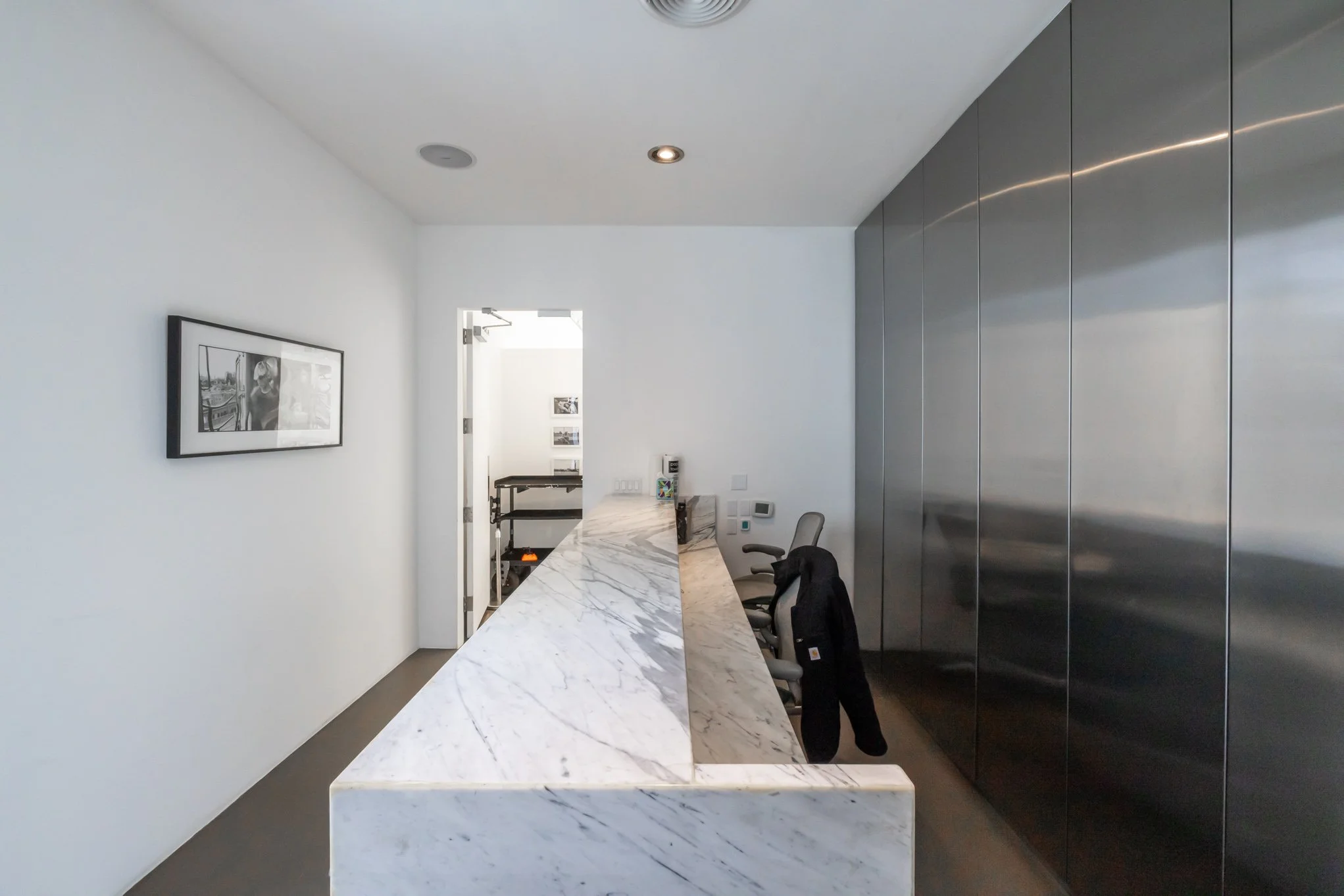 Modern office reception area with a marble counter, gray chairs, black suit jacket hanging, white walls, and stainless steel storage cabinets.