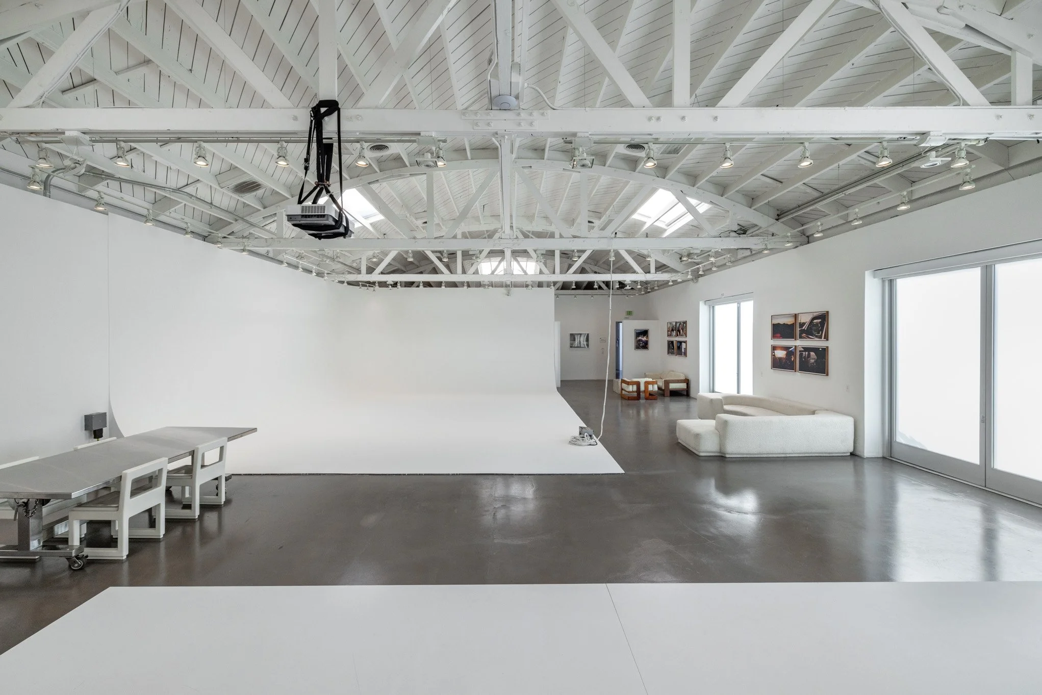 An empty art studio with a large white curved photography backdrop, white walls with framed photographs, a white couch, and a white table with chairs on a dark polished concrete floor. The ceiling features white exposed beams and skylights, with stud