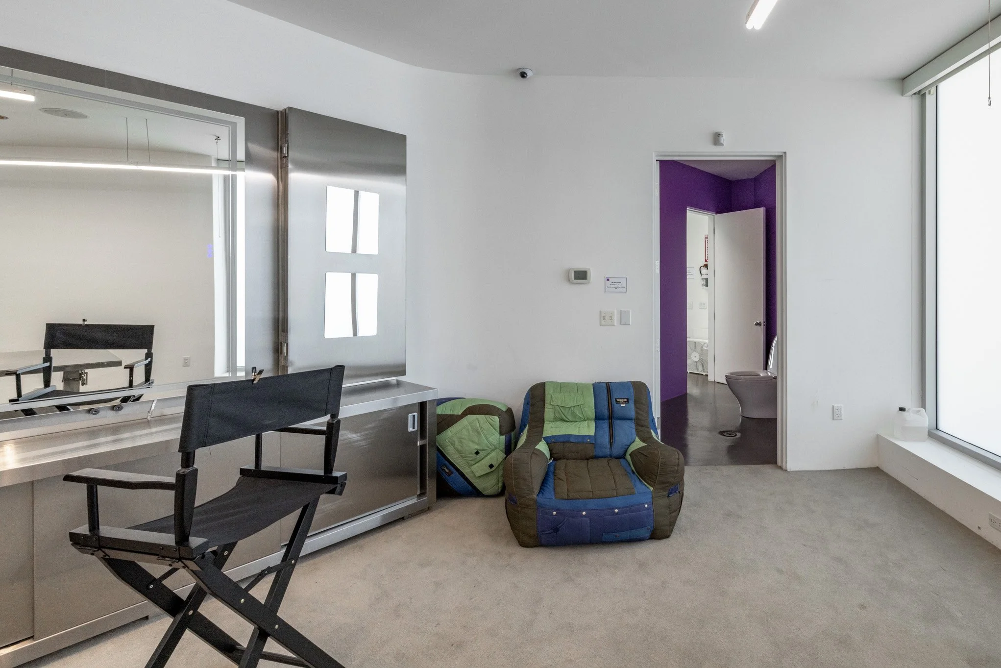 Interior of a room with a black director's chair, a luggage cart, two green and blue backpacks, and a brown and blue stuffed armchair. There is a purple accent wall leading to a bathroom with a visible toilet, and a large window on the right side wit