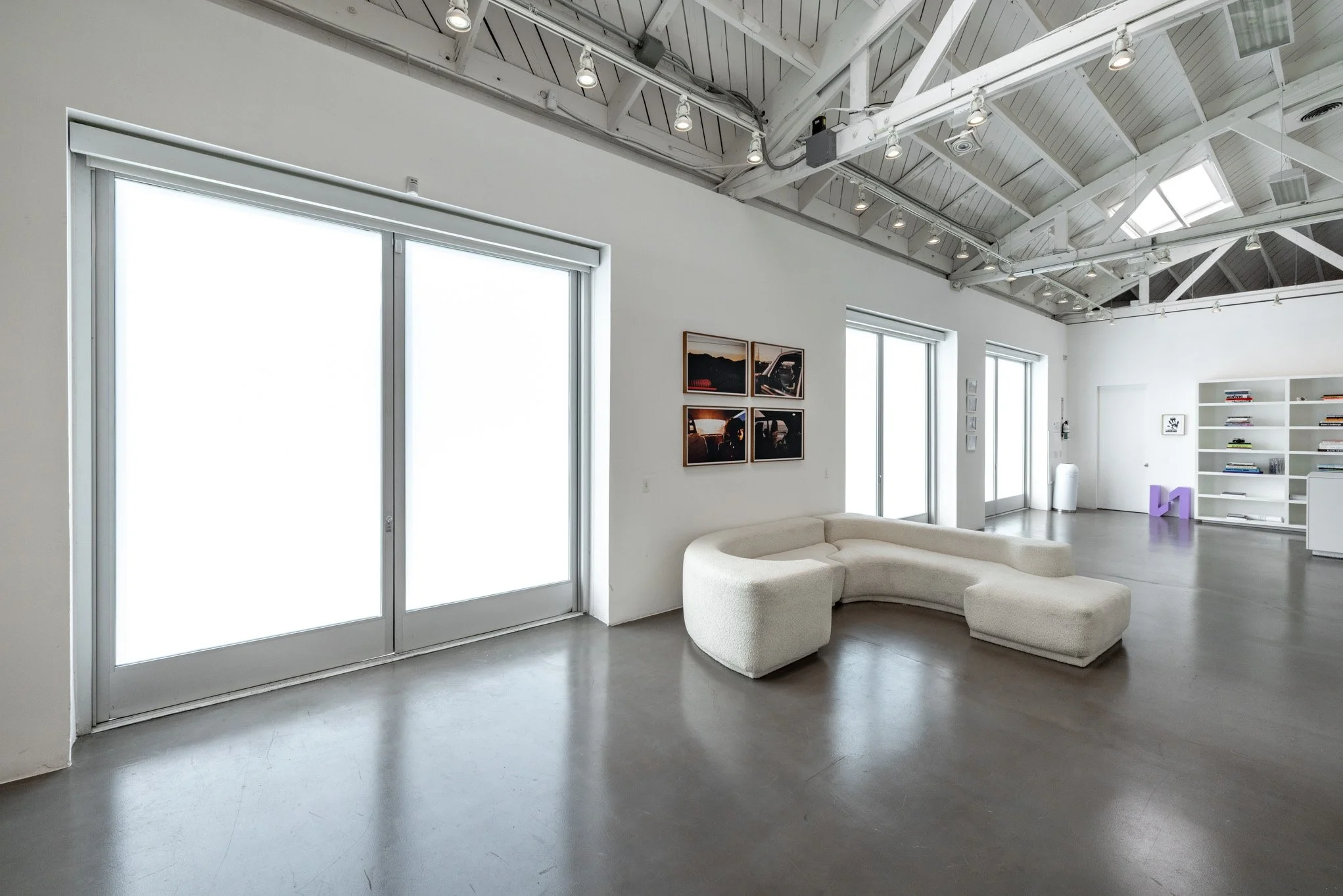 Modern, minimalist living room with large windows, a curved white sofa, and art on the wall inside a spacious, industrial-style building with white walls and high ceilings.