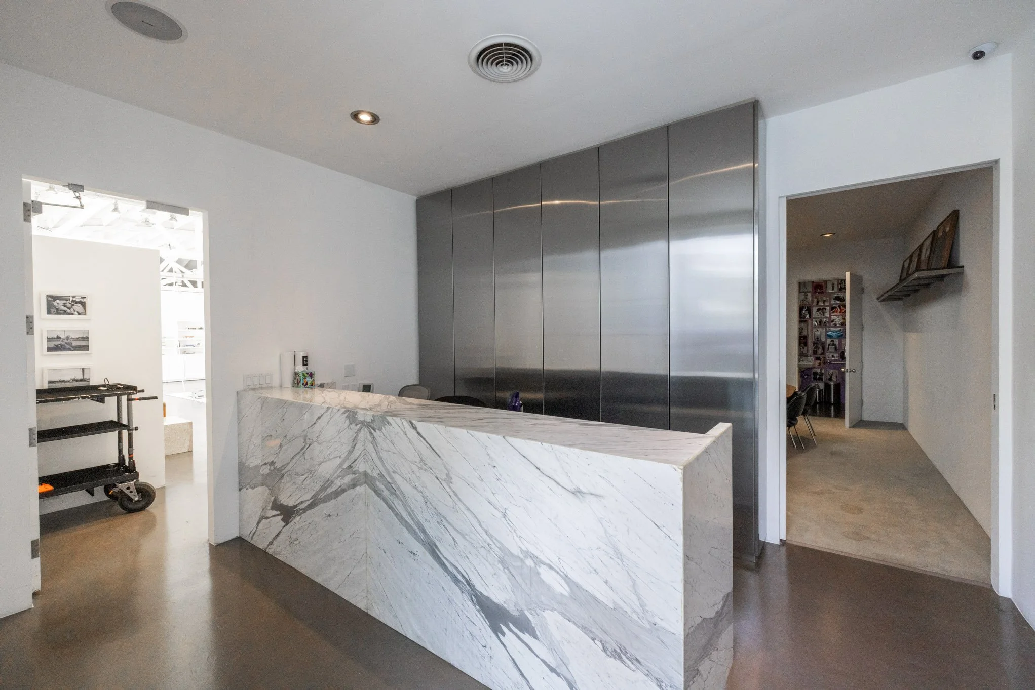 Modern reception area with a large white marble desk, stainless steel wall panels, and white walls. There is a doorway to the left leading to another room and a door to the right leading to a hallway with pictures on the wall.